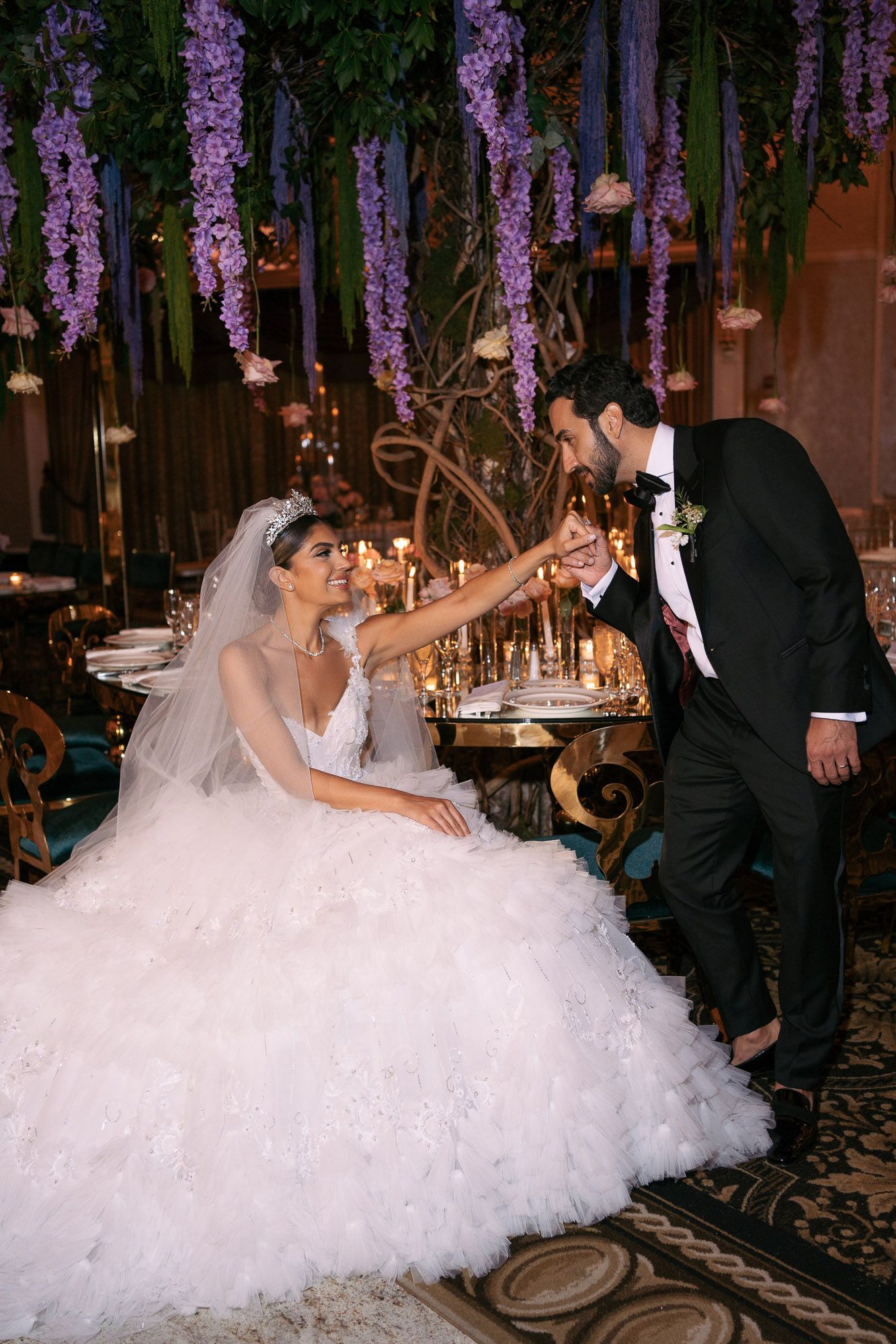 A bride in a voluminous white dress dances with her groom under hanging purple wisteria decorations at their reception.