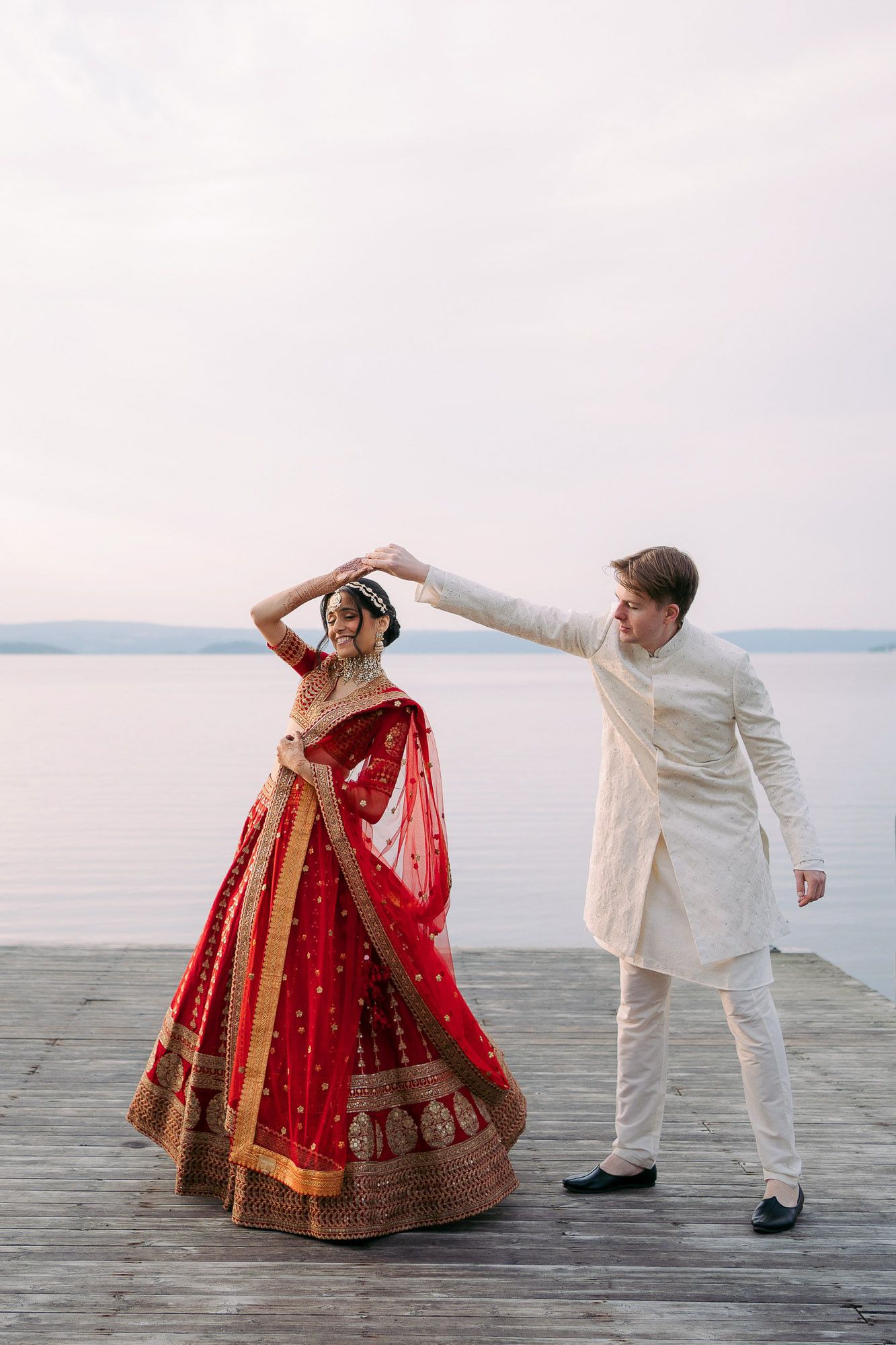 A couple in traditional wedding attire dance on a dock by the water, with the bride in a red and gold lehenga.