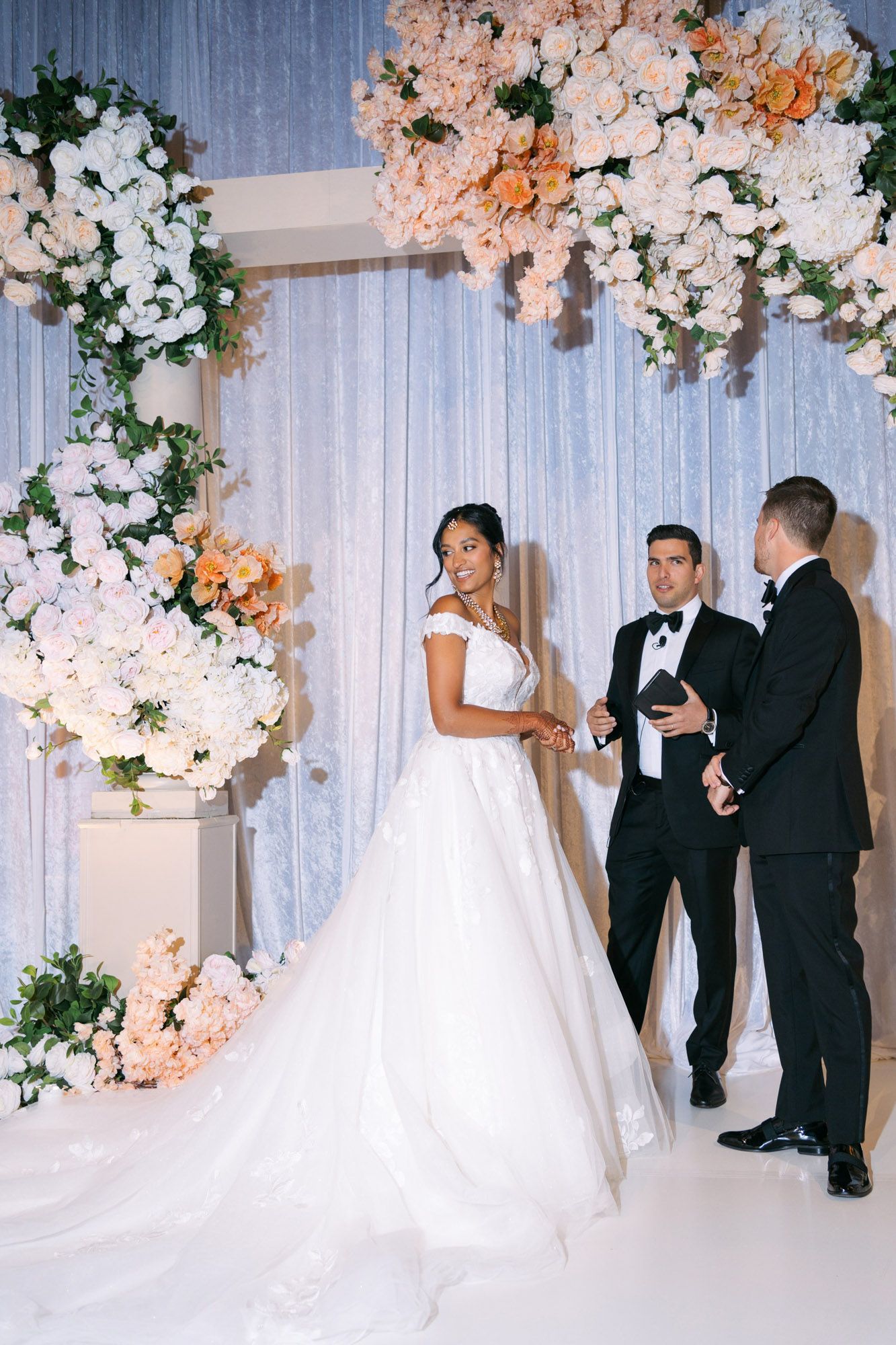 Elegant wedding ceremony setup with luxurious floral arch featuring white and peach roses against a silvery backdrop.