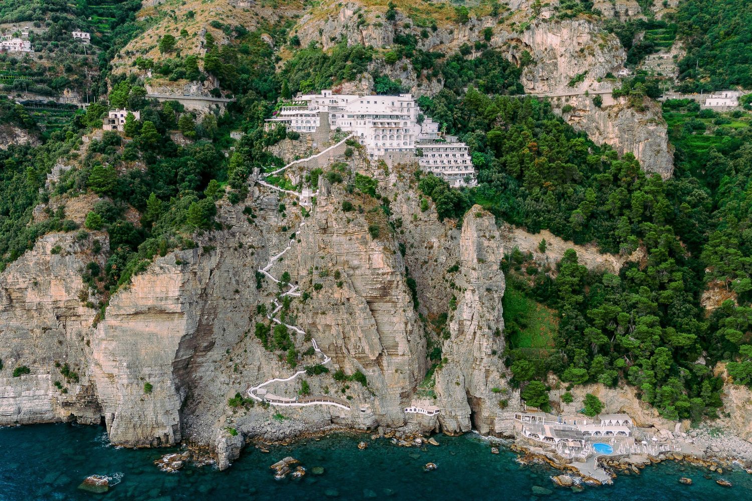 Aerial panoramic view of rocky coastline with boats anchored in turquoise Mediterranean waters near steep cliffs.