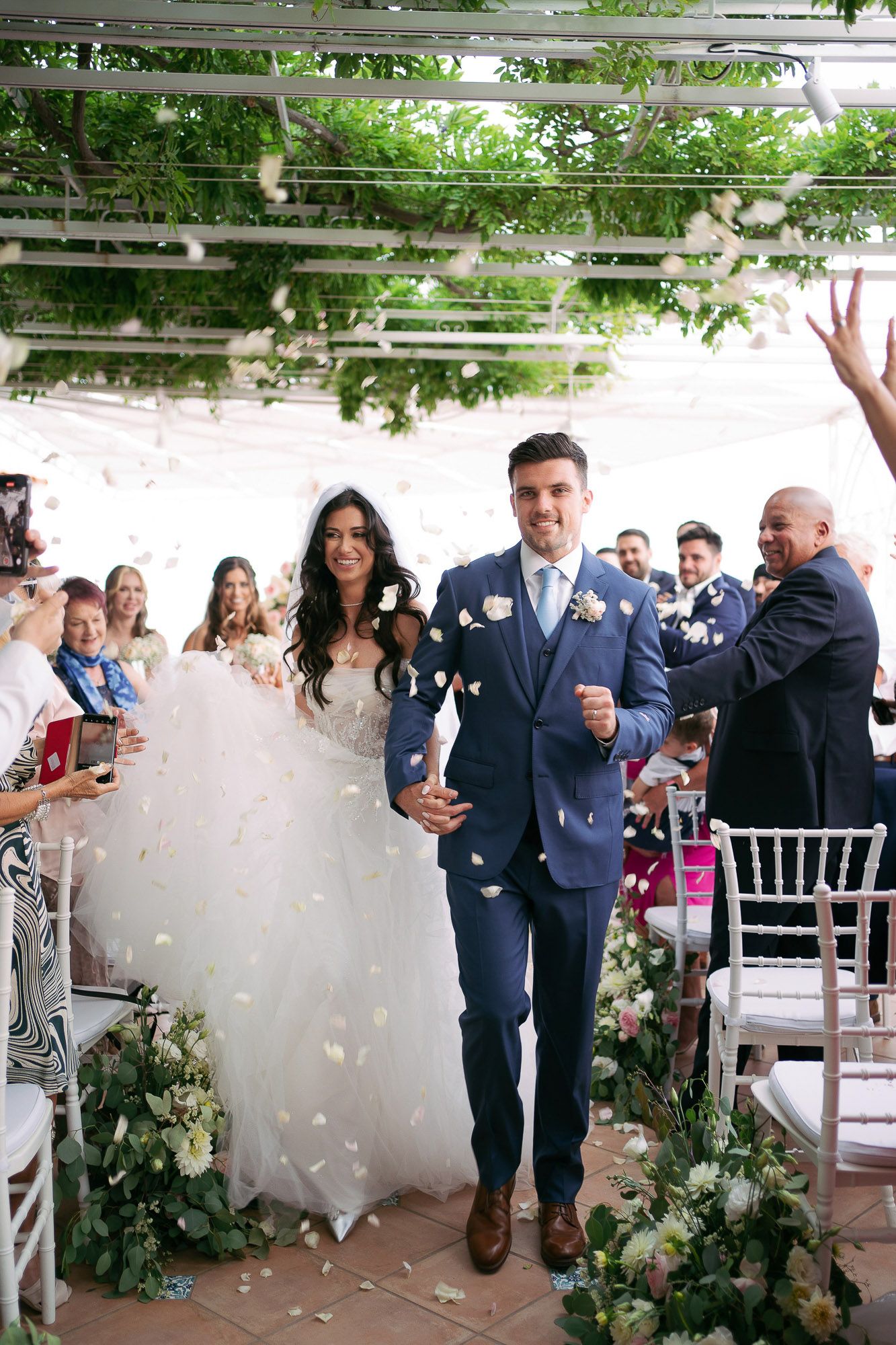 Wedding celebration with guests throwing flower petals at a garden venue decorated with greenery and white decor.