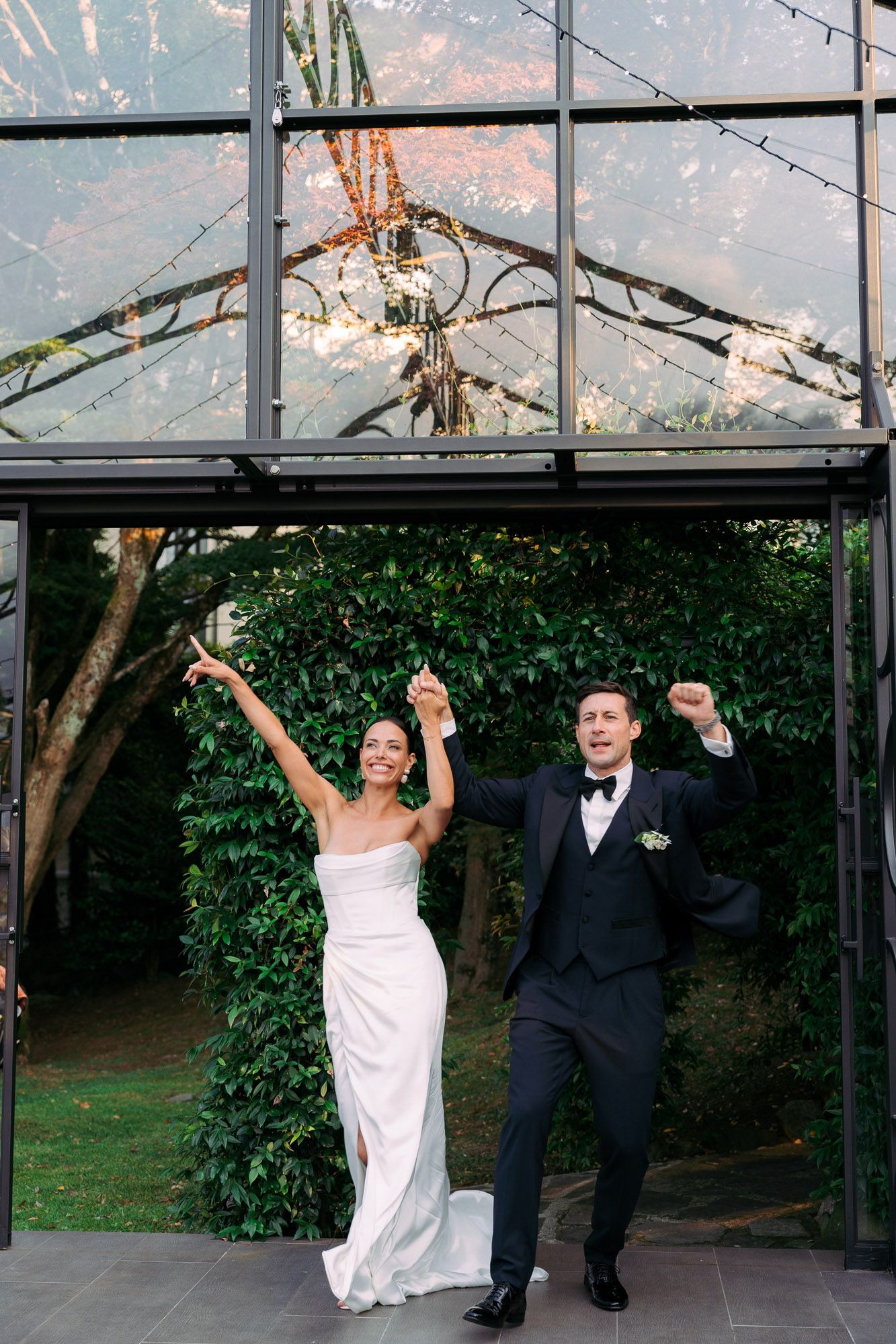 Newlywed couple celebrates joyfully while exiting a glass greenhouse venue decorated with string lights and greenery.