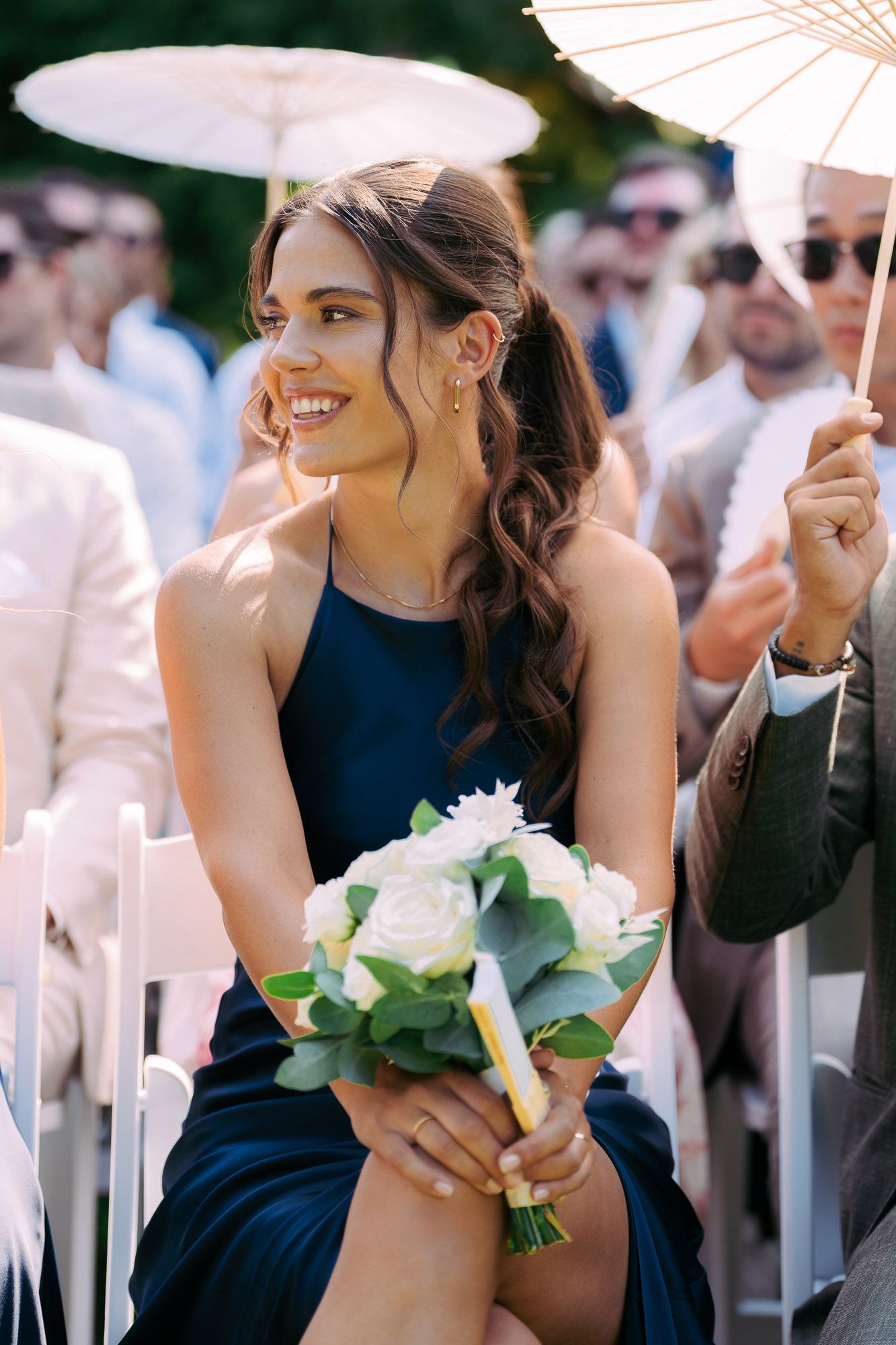 Bridesmaids in navy blue dresses hold white floral bouquets during an outdoor wedding ceremony.