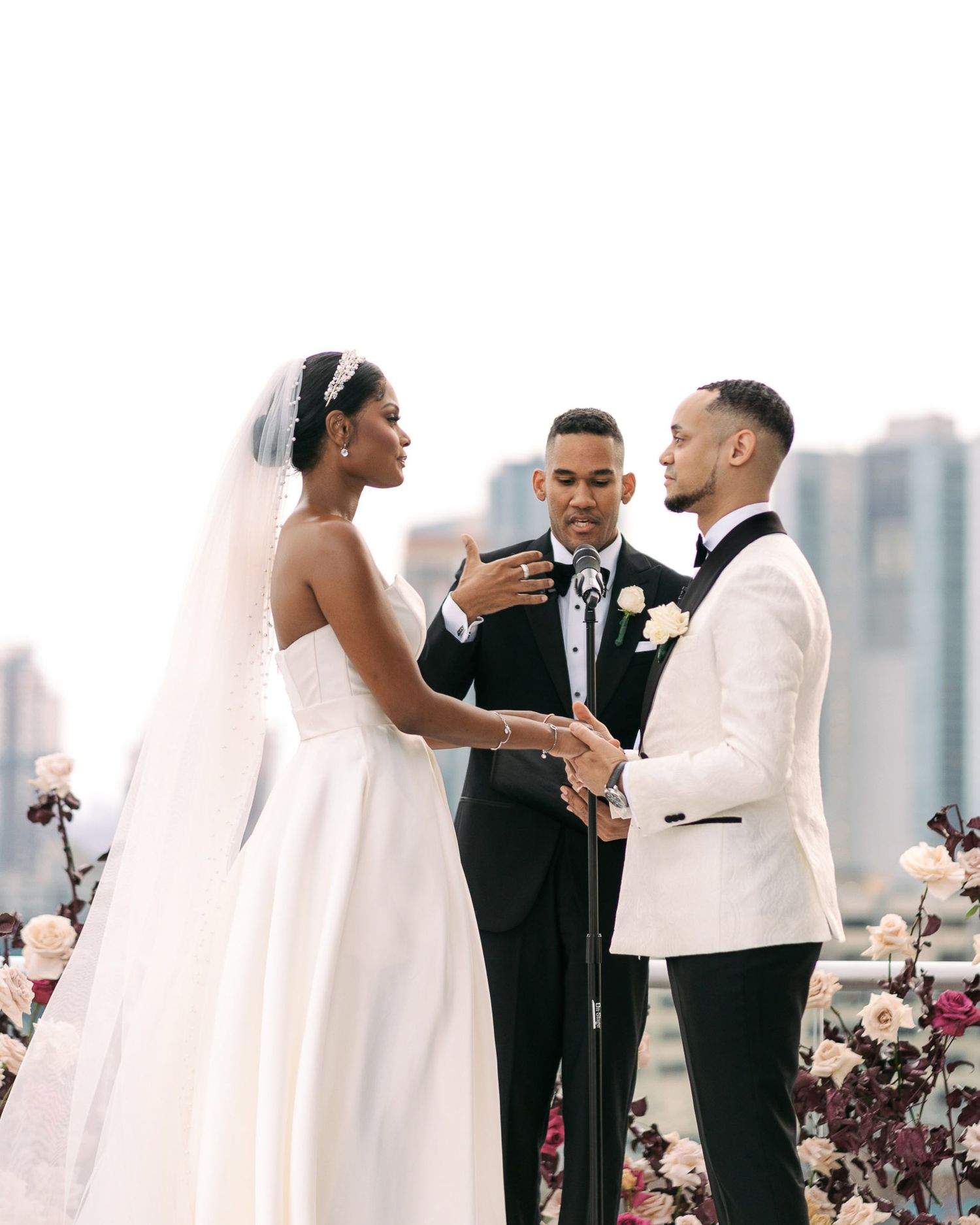 A wedding ceremony takes place on a rooftop with city skyline views and floral decorations in the background.