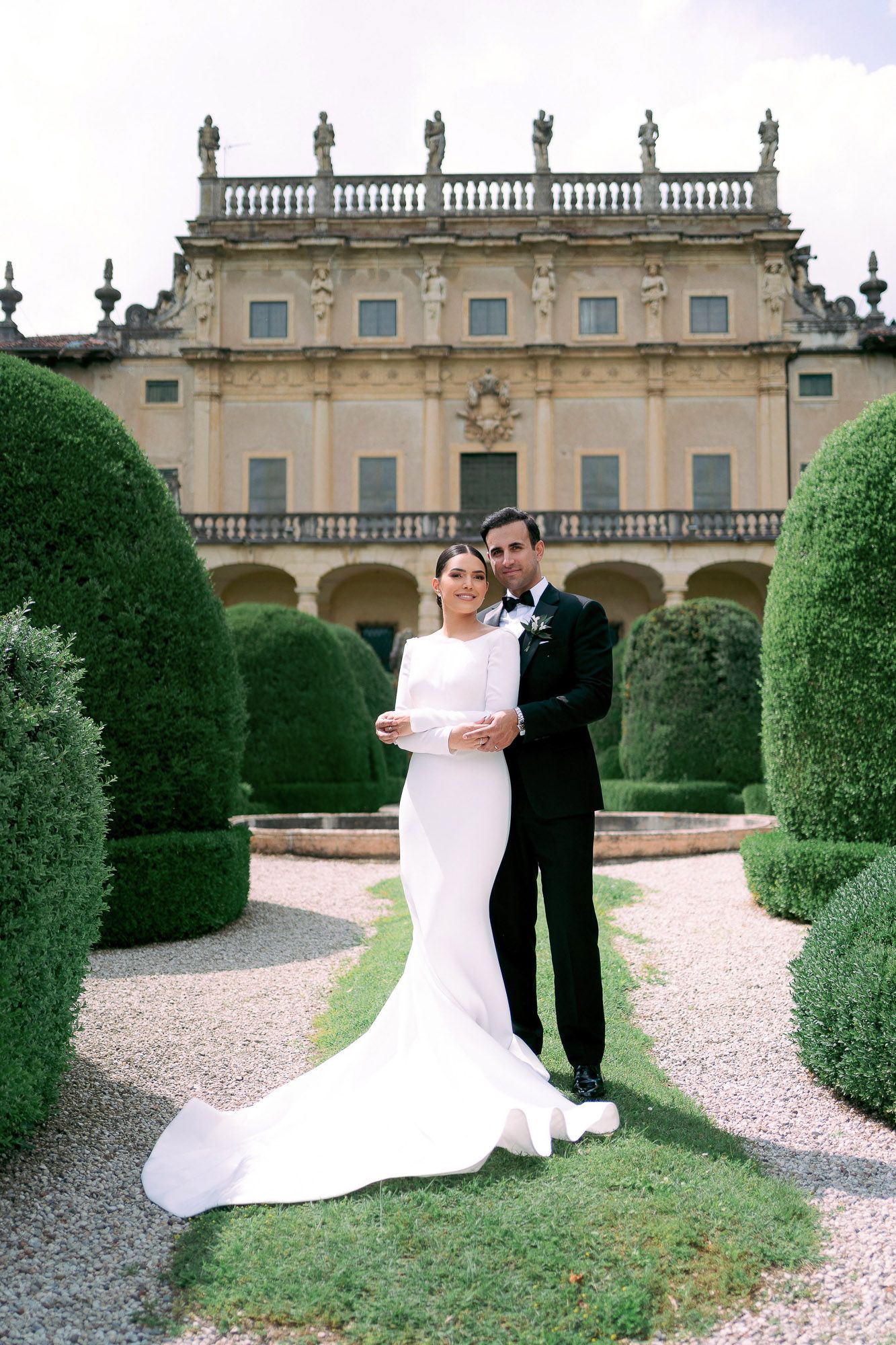 Elegant wedding couple poses in front of a historic stone mansion with manicured topiary gardens.