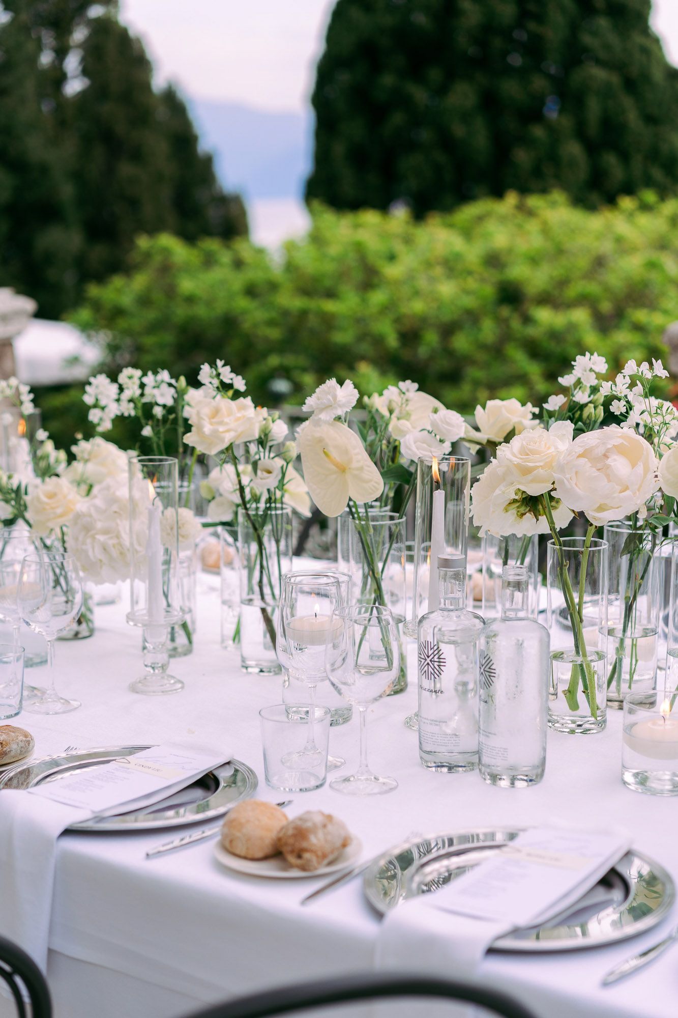 Elegant outdoor wedding table settings with white floral centerpieces in glass vases and crisp white linens.