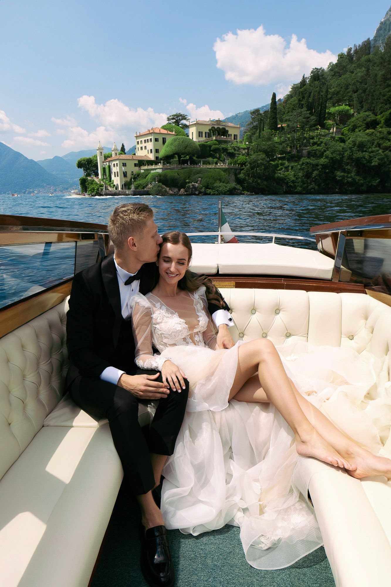 A couple relaxes on a luxury wooden boat on Lake Como with Italian villas and mountains in the background.