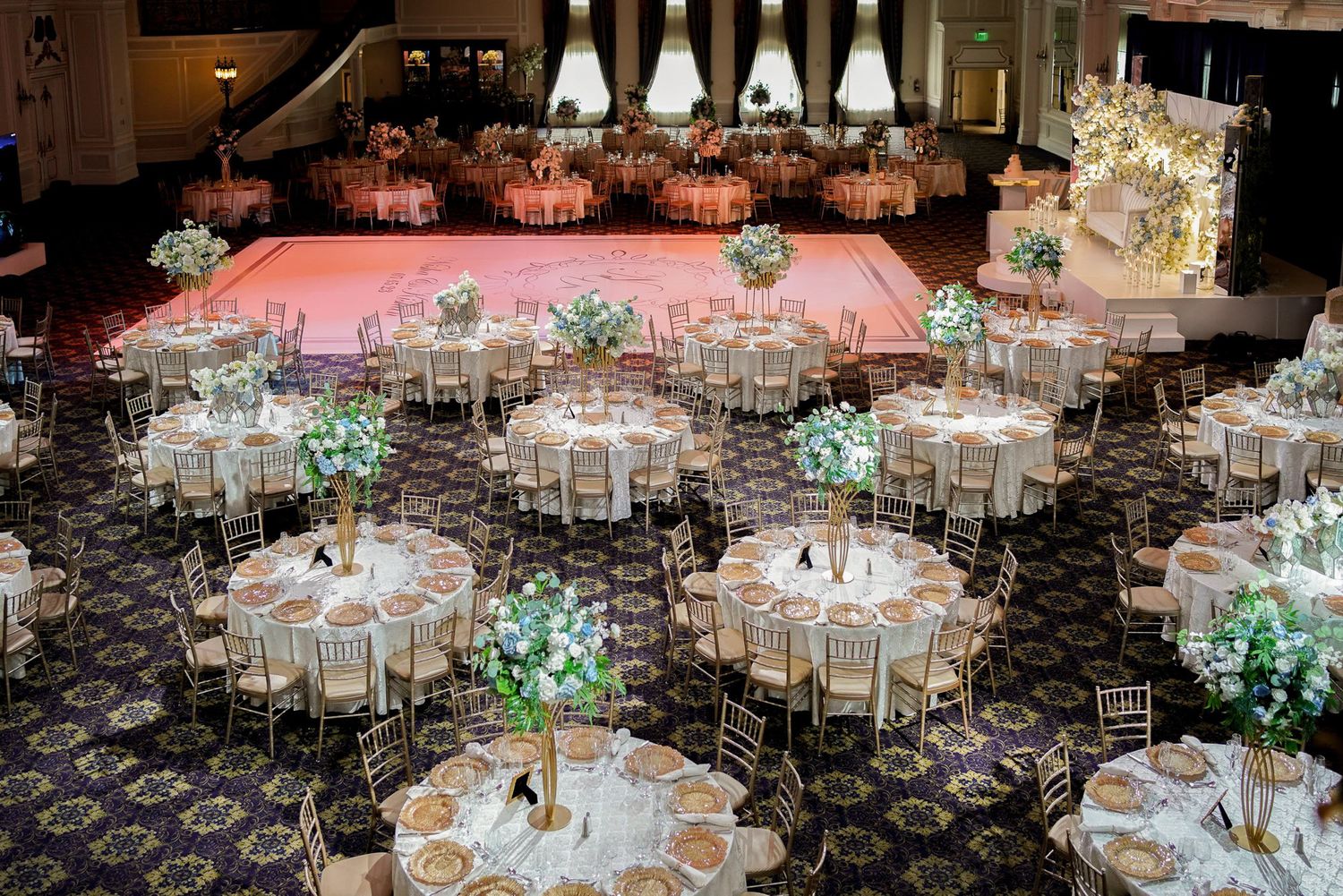 Elegant wedding reception ballroom with round tables, pink lighting, and floral centerpieces on patterned carpet.