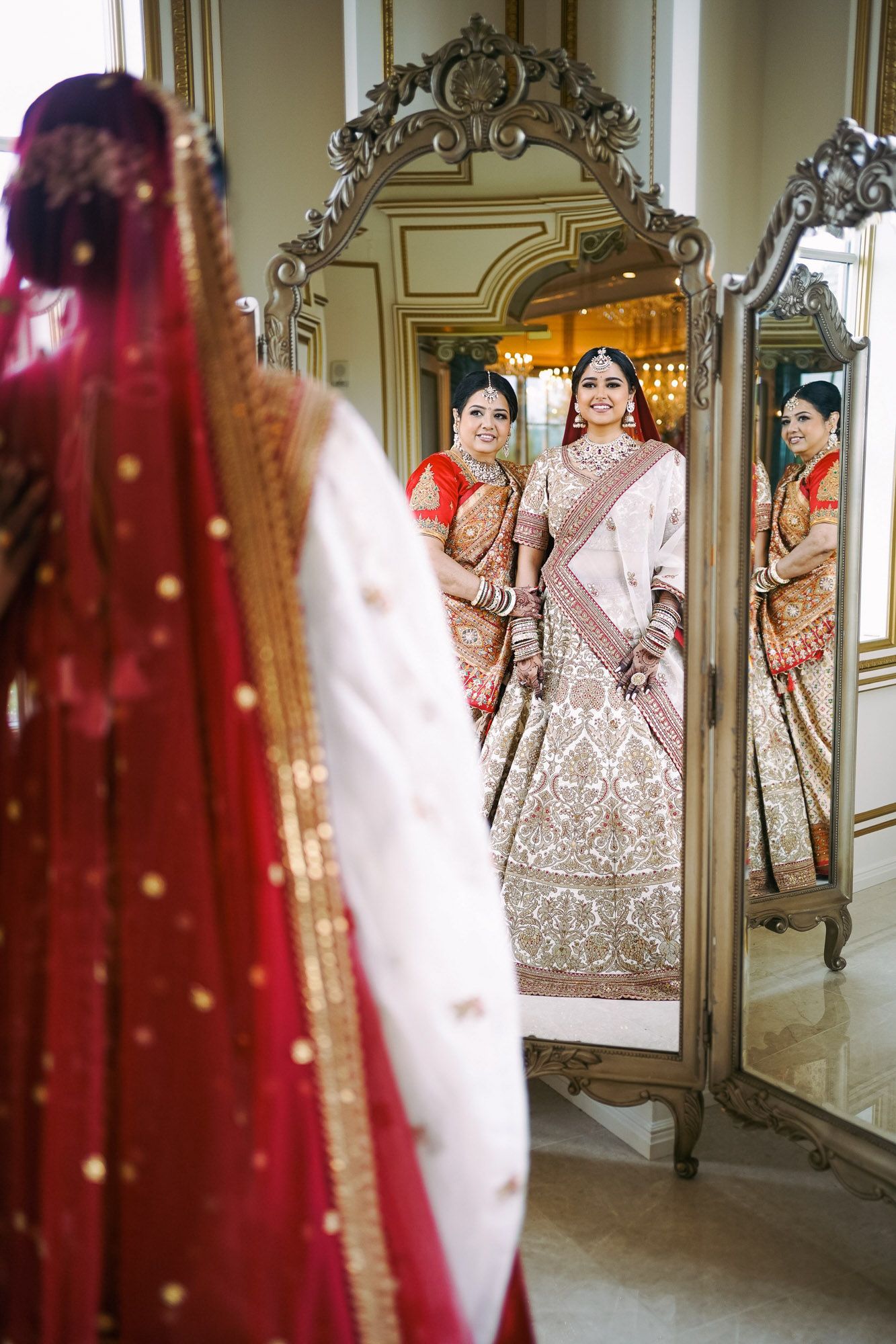 Elegant Indian bride in an ornate white and pink lehenga poses in front of decorative mirrors in an opulent hallway.