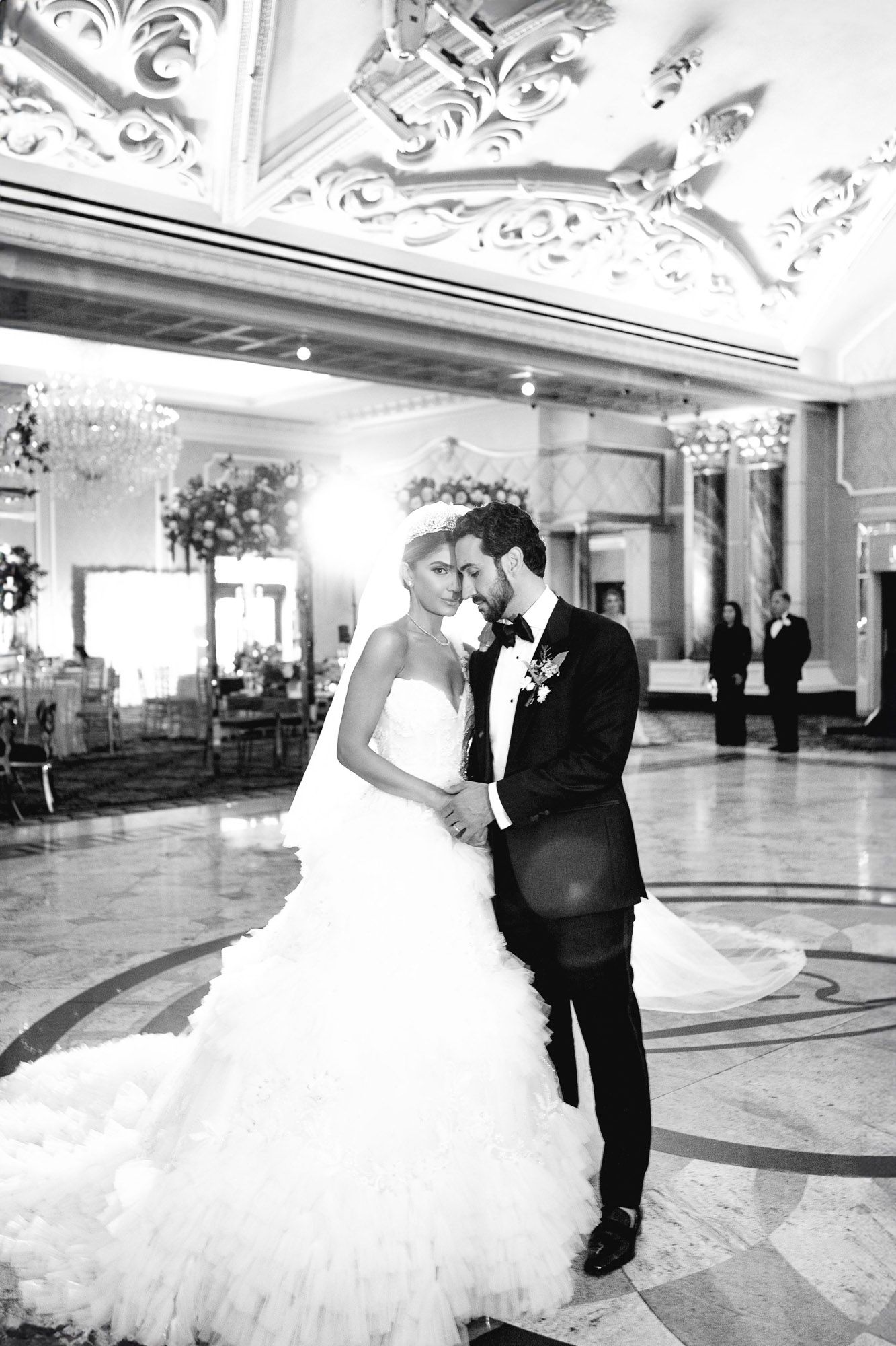Elegant couple shares their first dance in a luxurious ballroom with ornate ceiling details and marble floors.