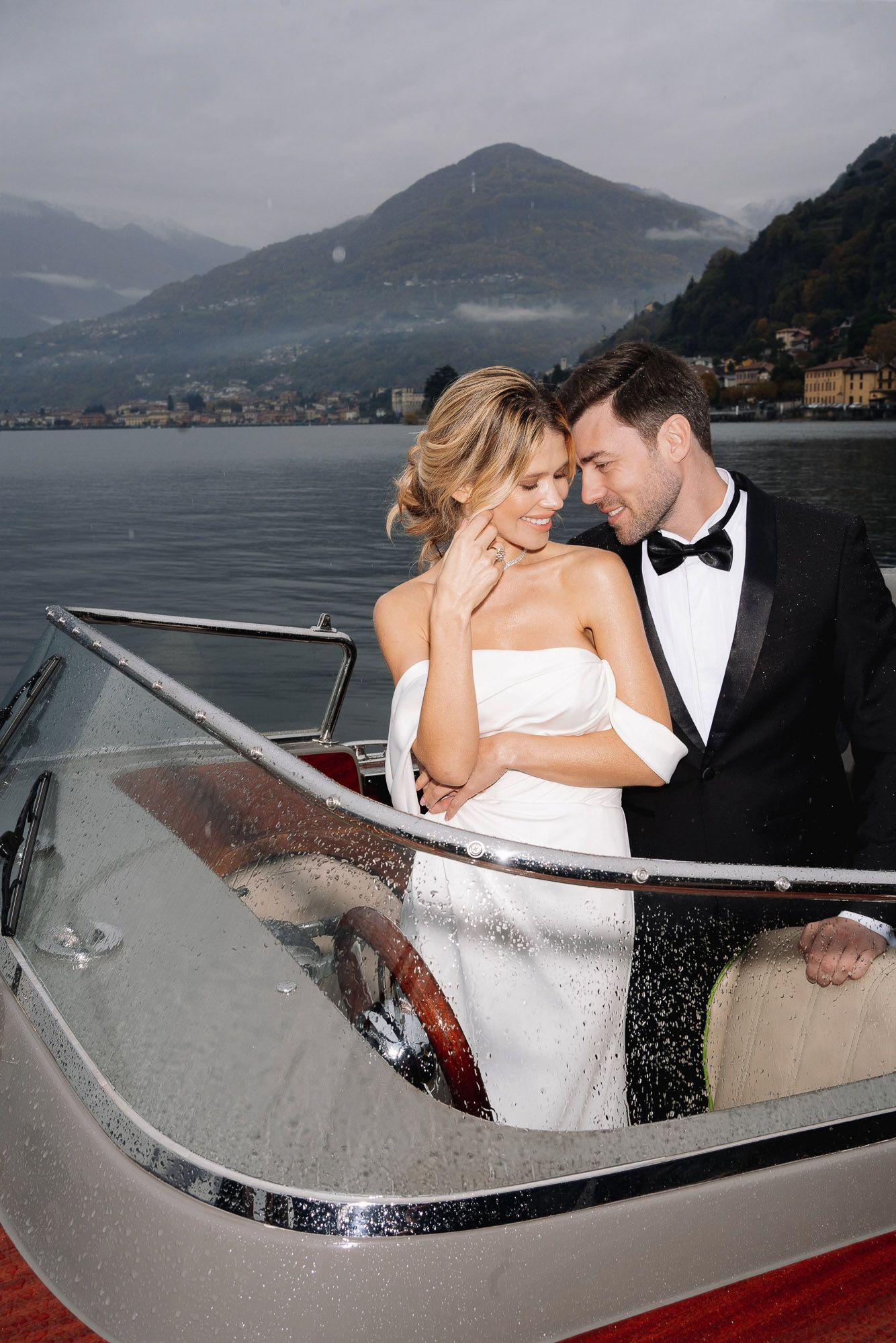 A couple shares romantic moments in a vintage boat on a scenic lake surrounded by mountains.