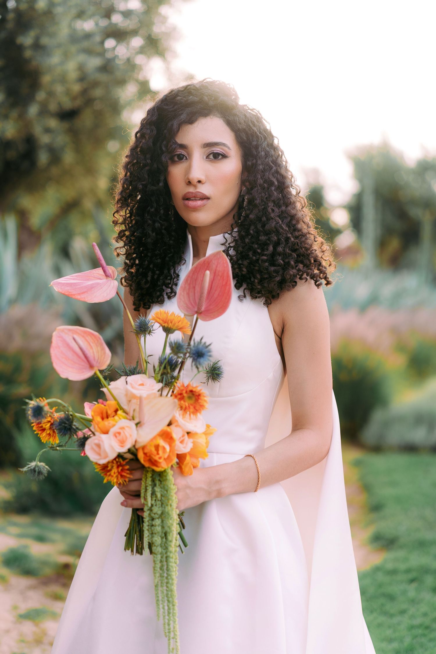 Bride in white dress holding vibrant orange and coral bouquet in garden setting.