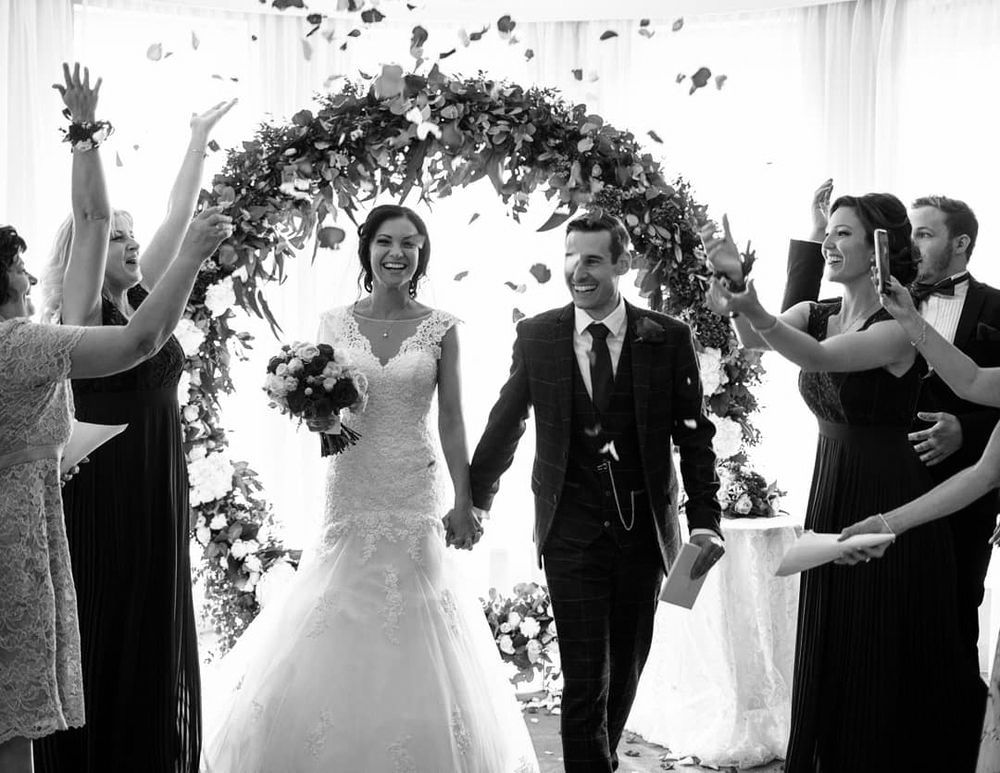 Black and white photo of wedding ceremony featuring bride and groom standing with wedding guests in an indoor venue.