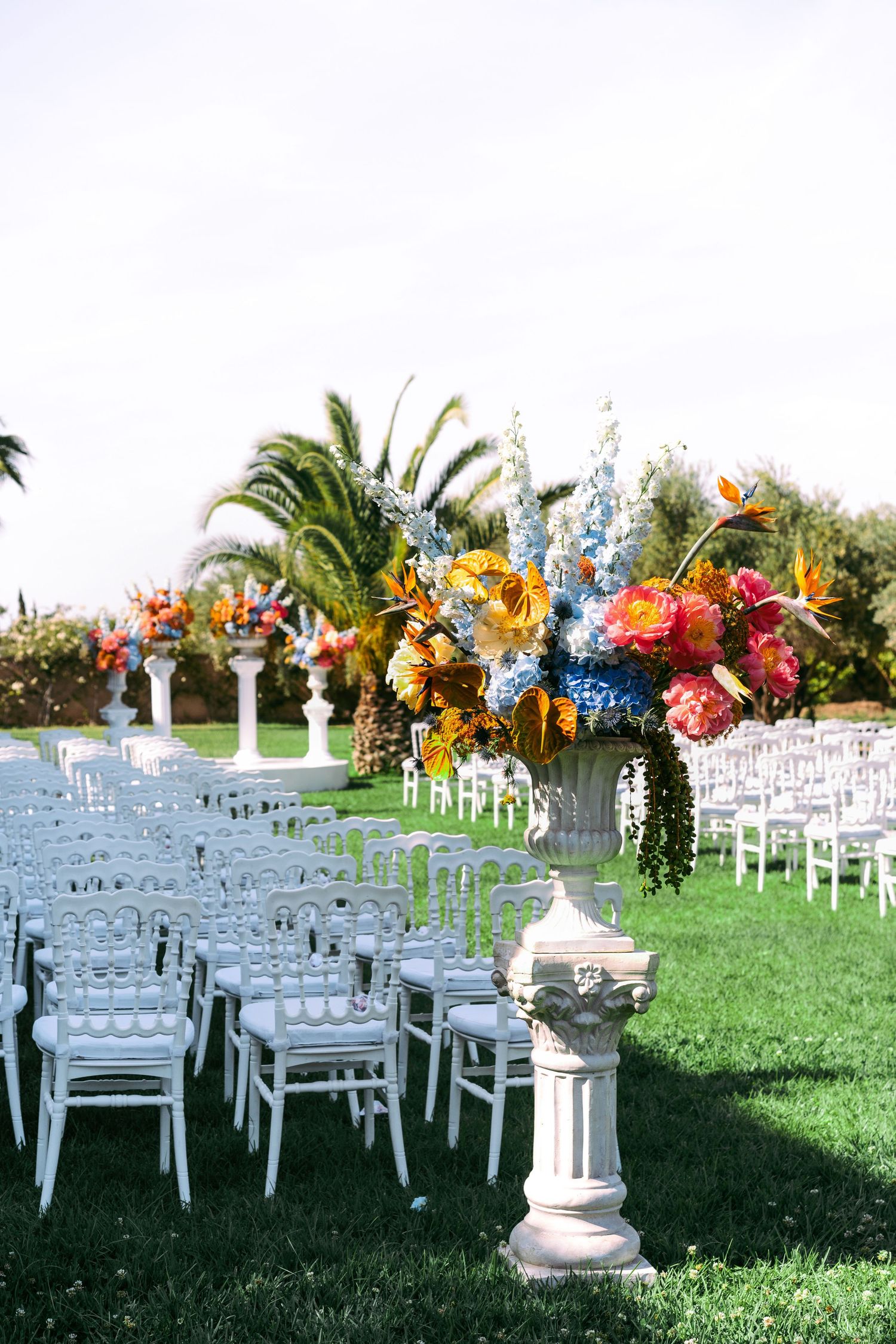 Outdoor wedding ceremony setup with white chairs and colorful floral arrangements under palm trees and blue sky.