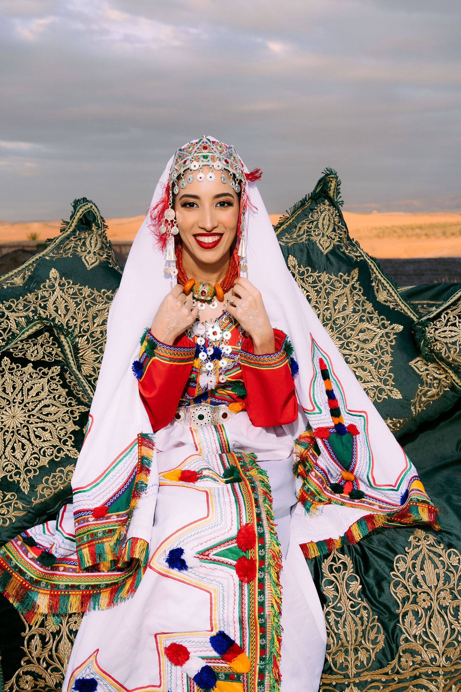 Colorful traditional Moroccan wedding attire featuring ornate white and red clothing with decorative embellishments against desert backdrop.