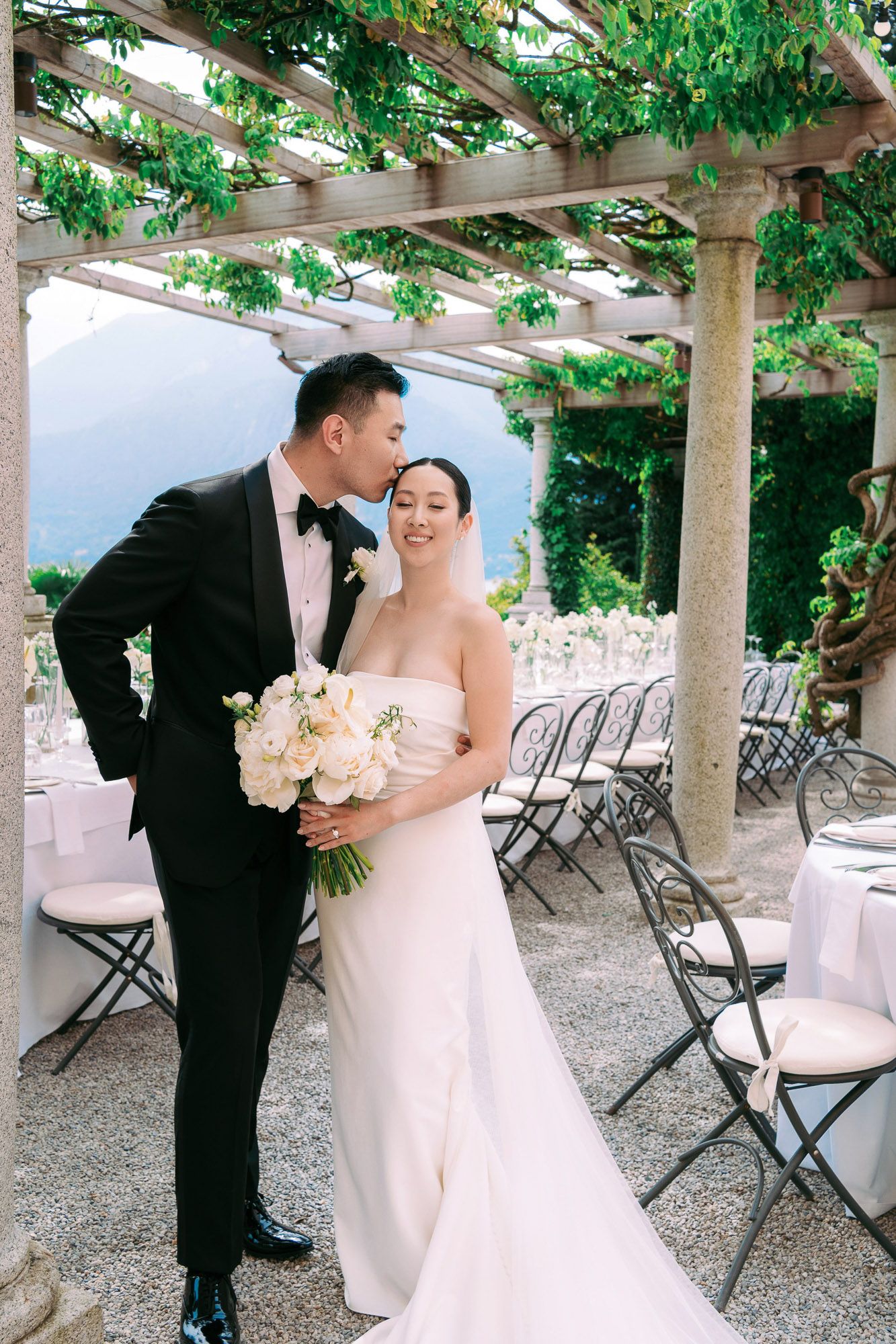 A romantic wedding couple stands beneath a vine-covered pergola with folding chairs arranged in the background.