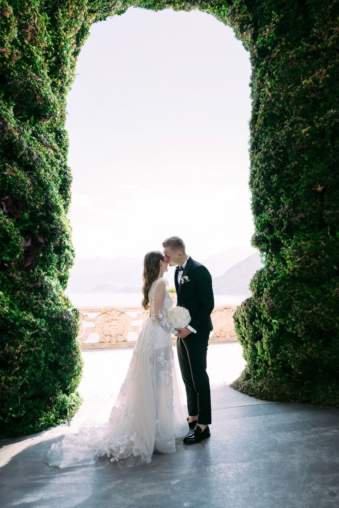 A romantic wedding couple shares an intimate moment framed by lush green cypress trees in an outdoor setting.