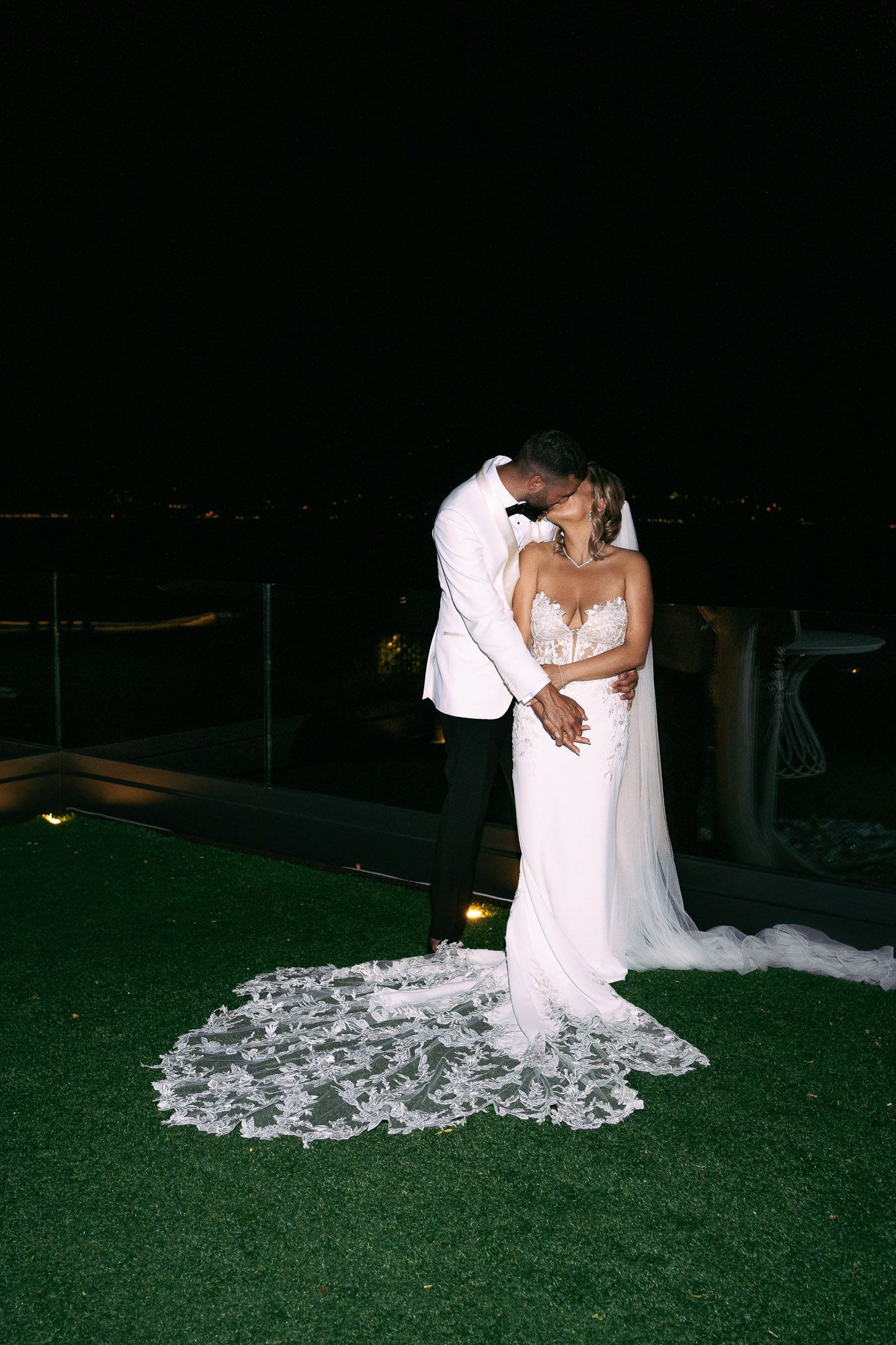 A couple in wedding attire embraces on a dark lawn with a flowing white lace train trailing behind them.