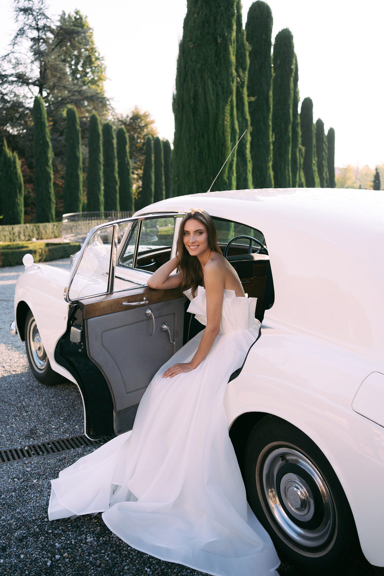 Elegant white wedding dress flows out of classic car door with tall cypress trees in background.