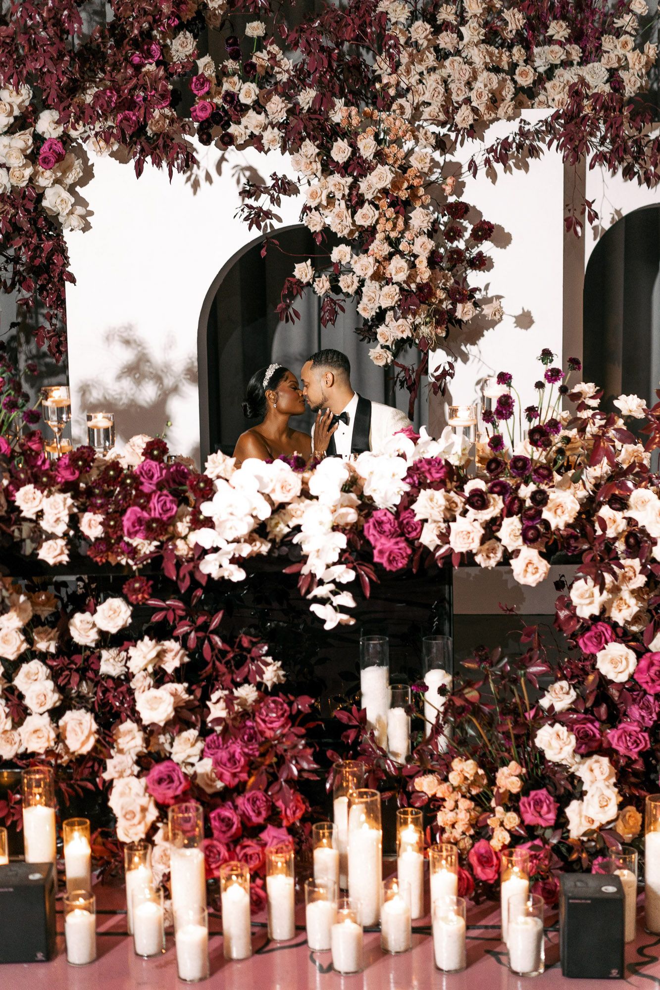 Elegant wedding decor features white pillar candles and cascading pink and white floral arrangements against a dark backdrop.