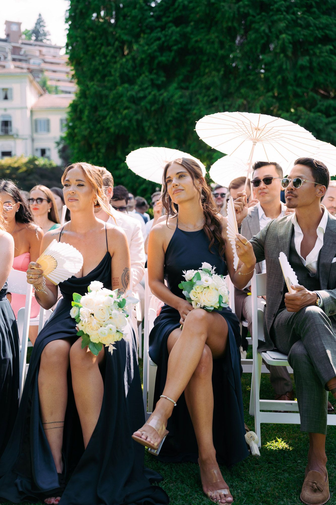 Wedding party members in formal black attire pose playfully in a line outdoors during a sunny celebration.