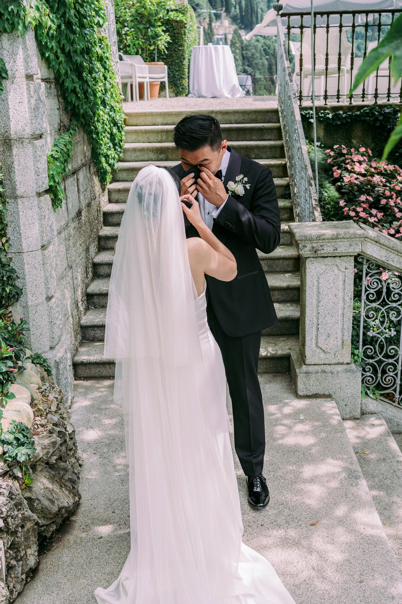 A couple in wedding attire shares an intimate moment on stone steps surrounded by greenery and pink flowers.