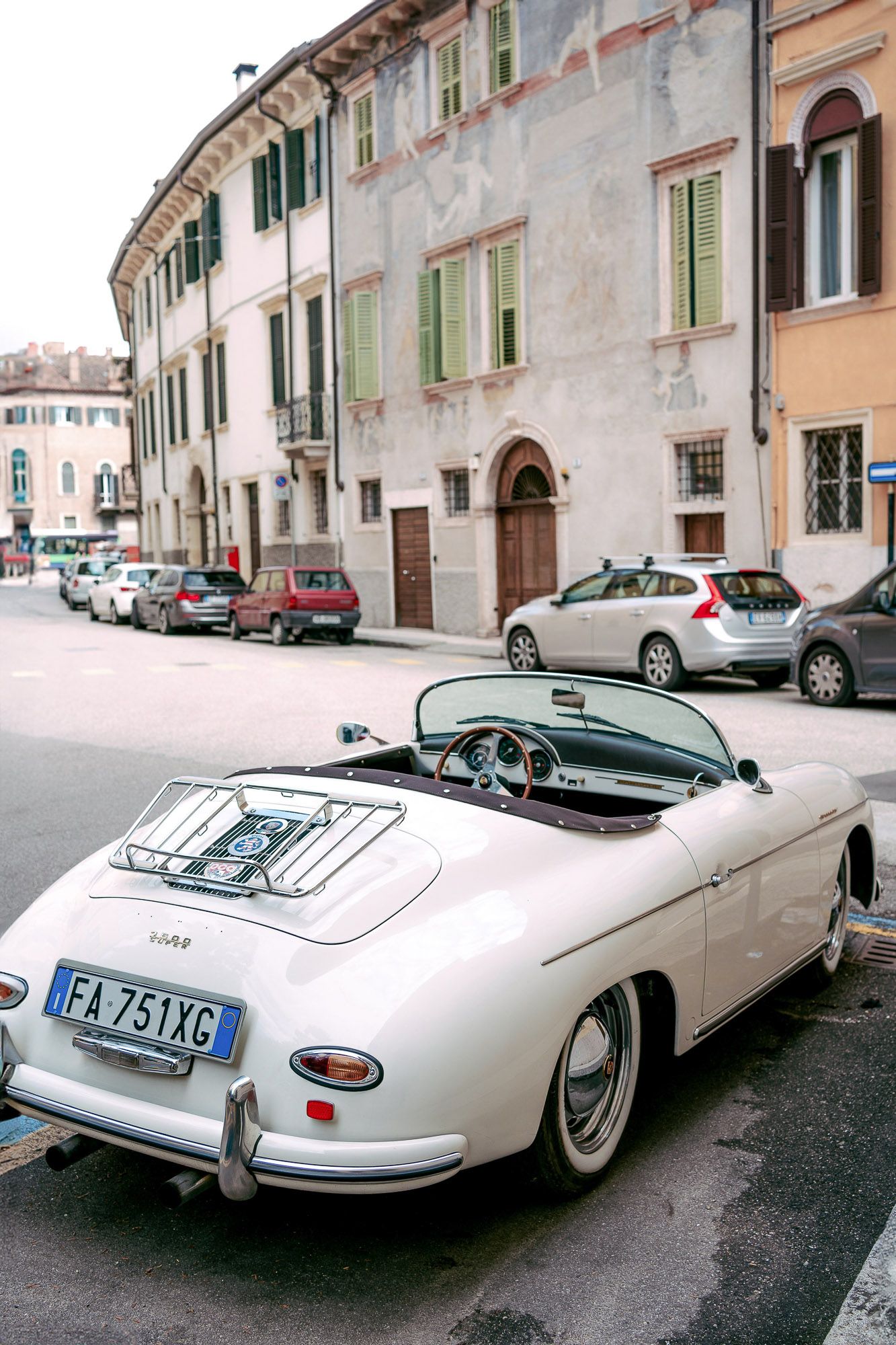 A classic white Porsche 356 convertible parked on a cobblestone street in front of historic European buildings.