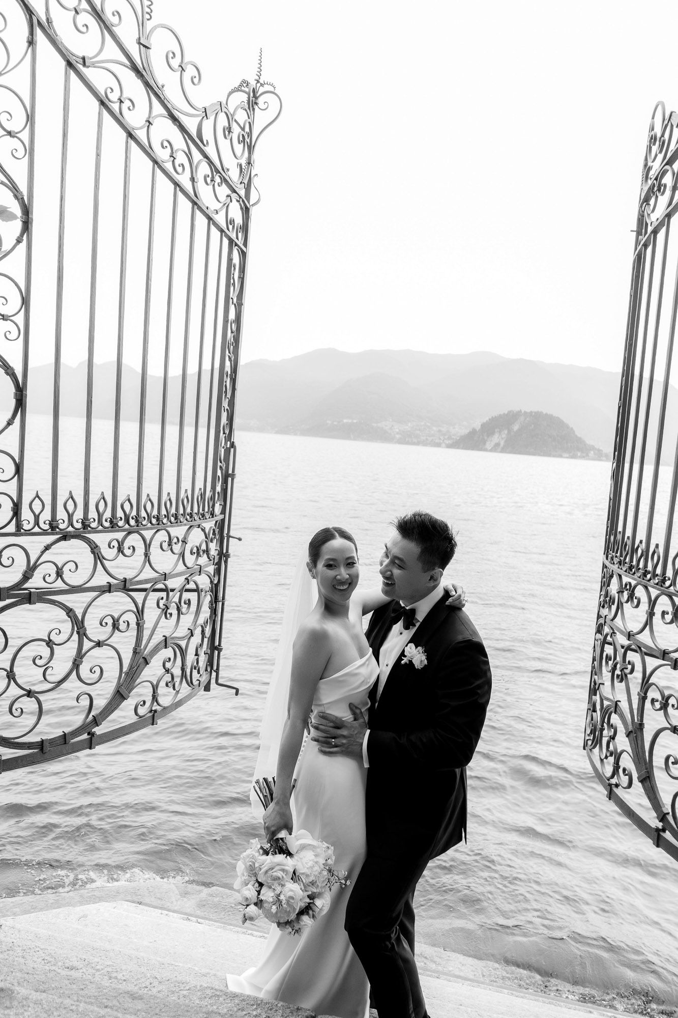 Elegant wedding couple poses in front of ornate gates overlooking a serene lake with mountains in the distance.