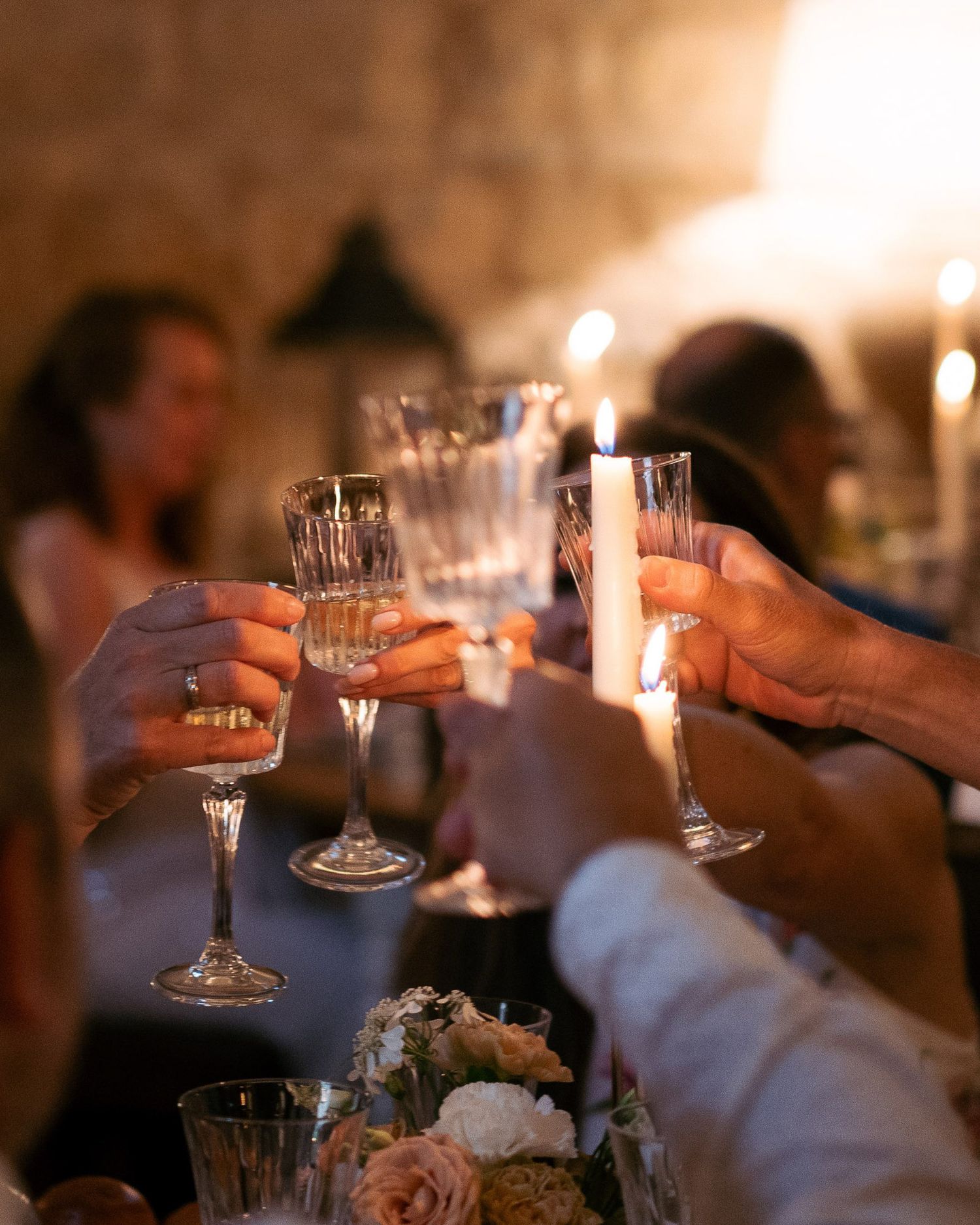 Hands clink champagne glasses together in a dimly lit celebration setting with candlelight and floral centerpieces.