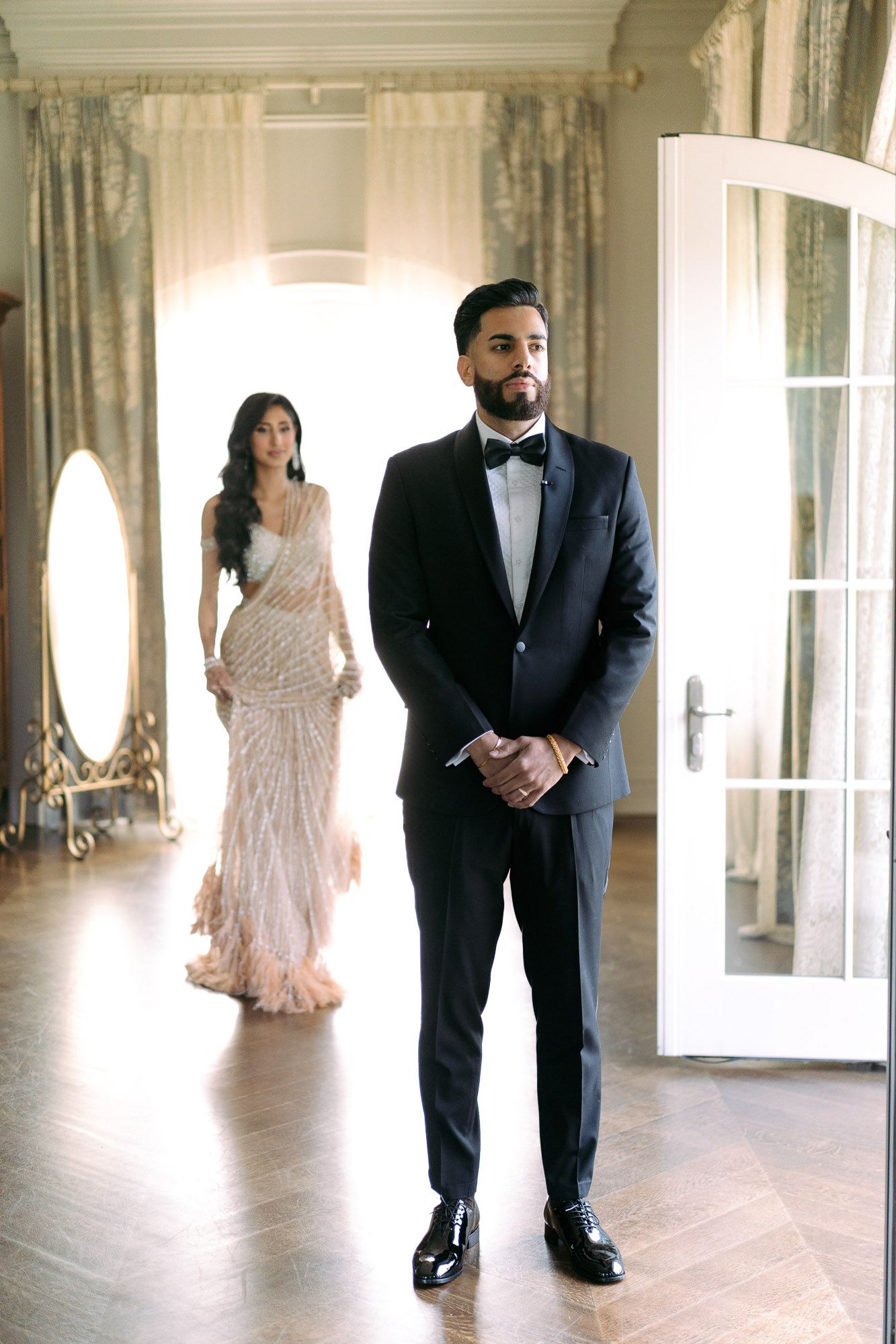 Elegant couple in formal attire poses for wedding photos in a bright sunlit venue with French doors and white walls.