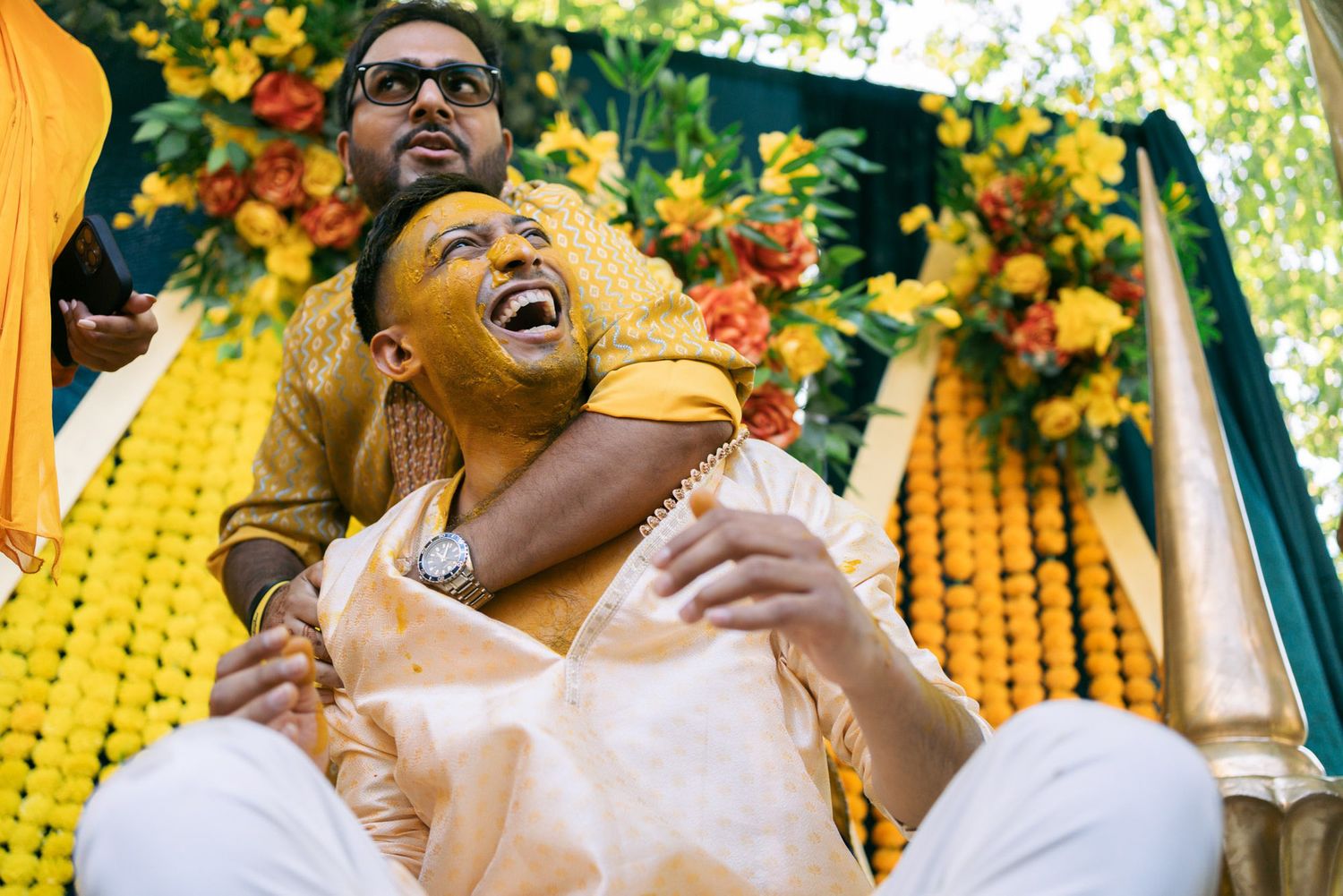 Joyful couple embraces and laughs together on swing decorated with vibrant yellow marigold flowers at outdoor celebration.