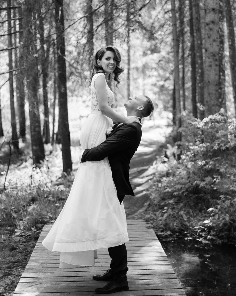 A romantic black and white wedding photo of a couple embracing on a wooden walkway in a misty forest setting.