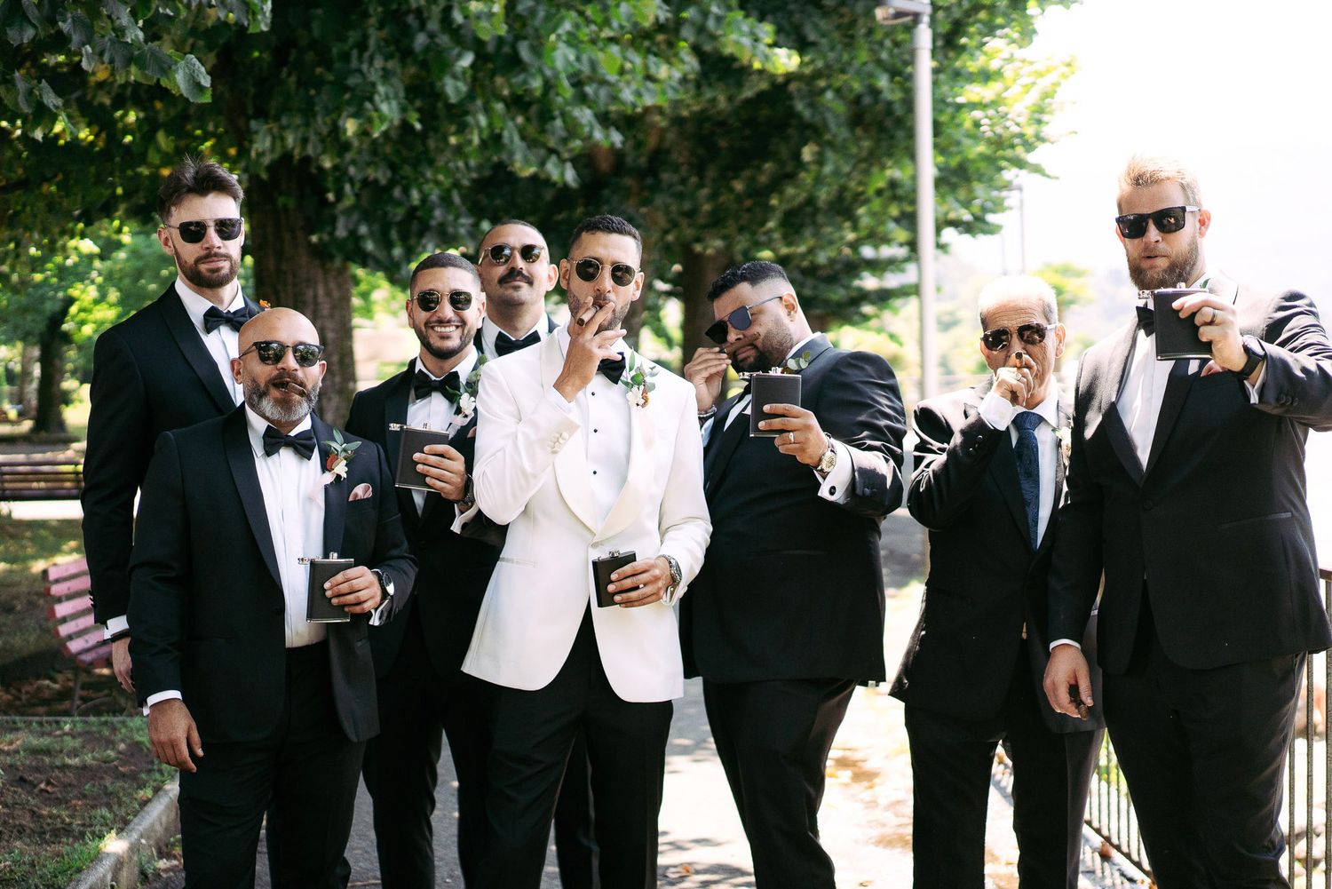 A wedding party in formal black and white attire poses together outdoors while holding drinks and wearing sunglasses.