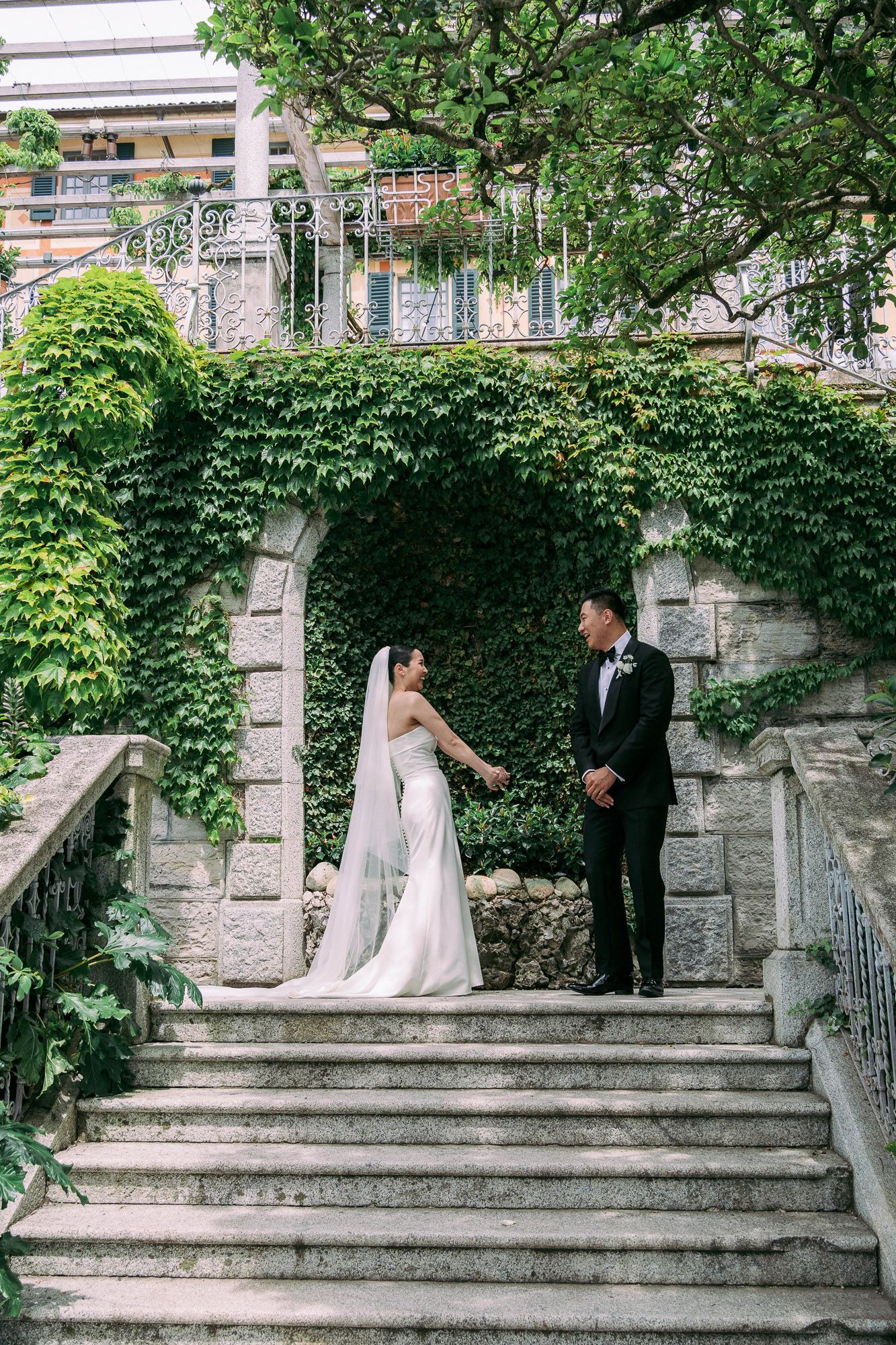Couple in formal wedding attire standing on stone steps surrounded by lush green foliage at outdoor venue.