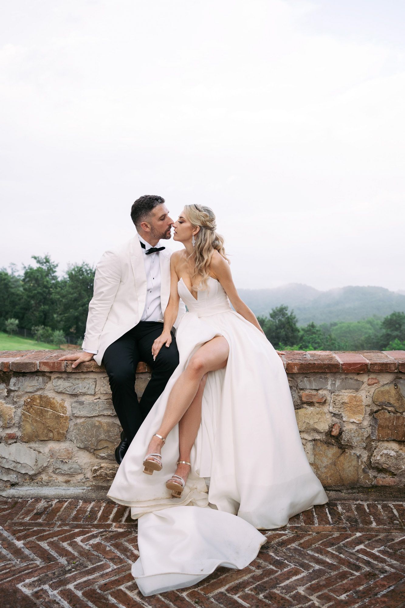 Romantic wedding couple sharing intimate kisses on a rustic stone wall overlooking mountain scenery.