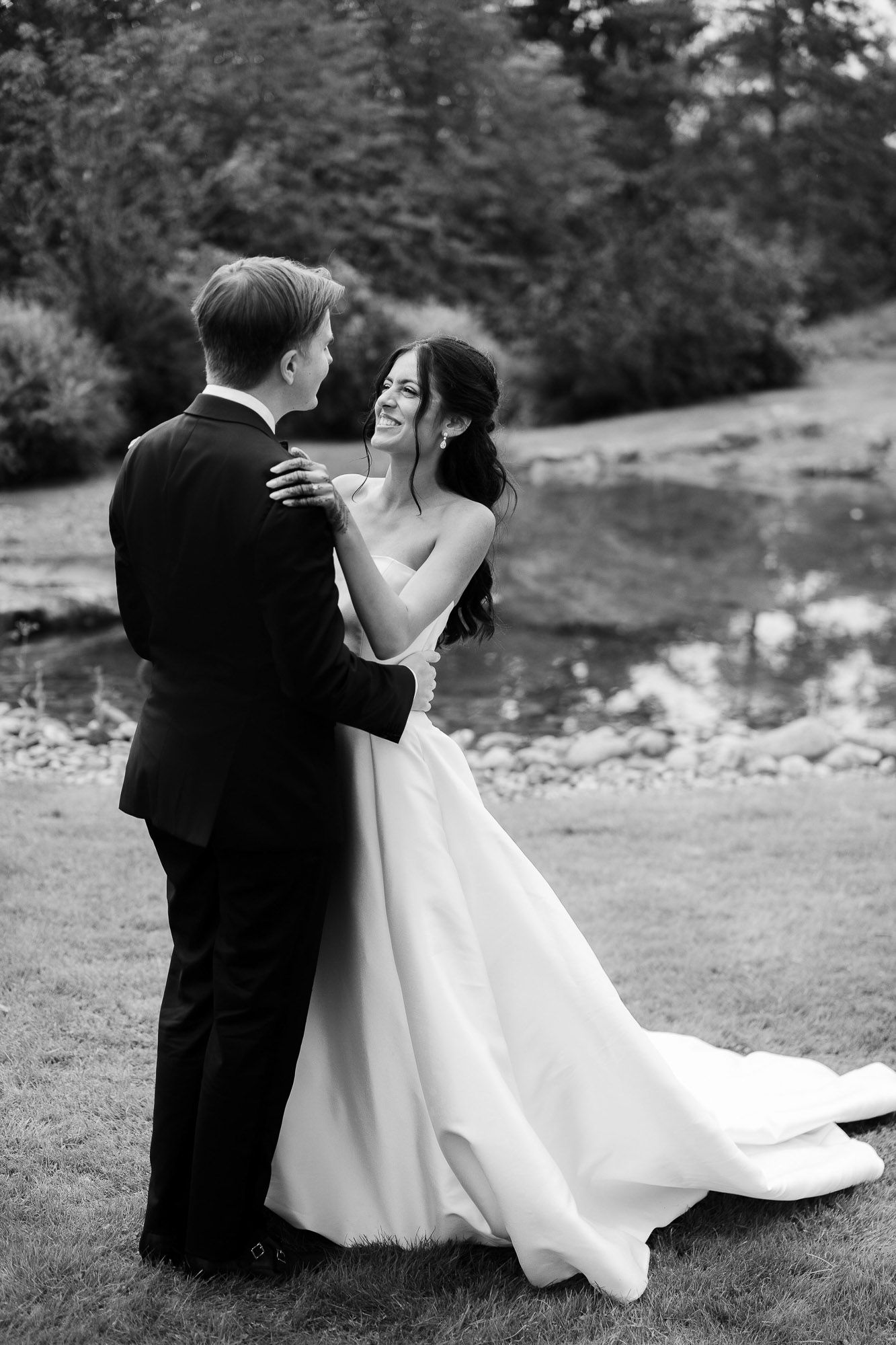 A couple in wedding attire shares a romantic moment together on grassy terrain near a river in black and white.