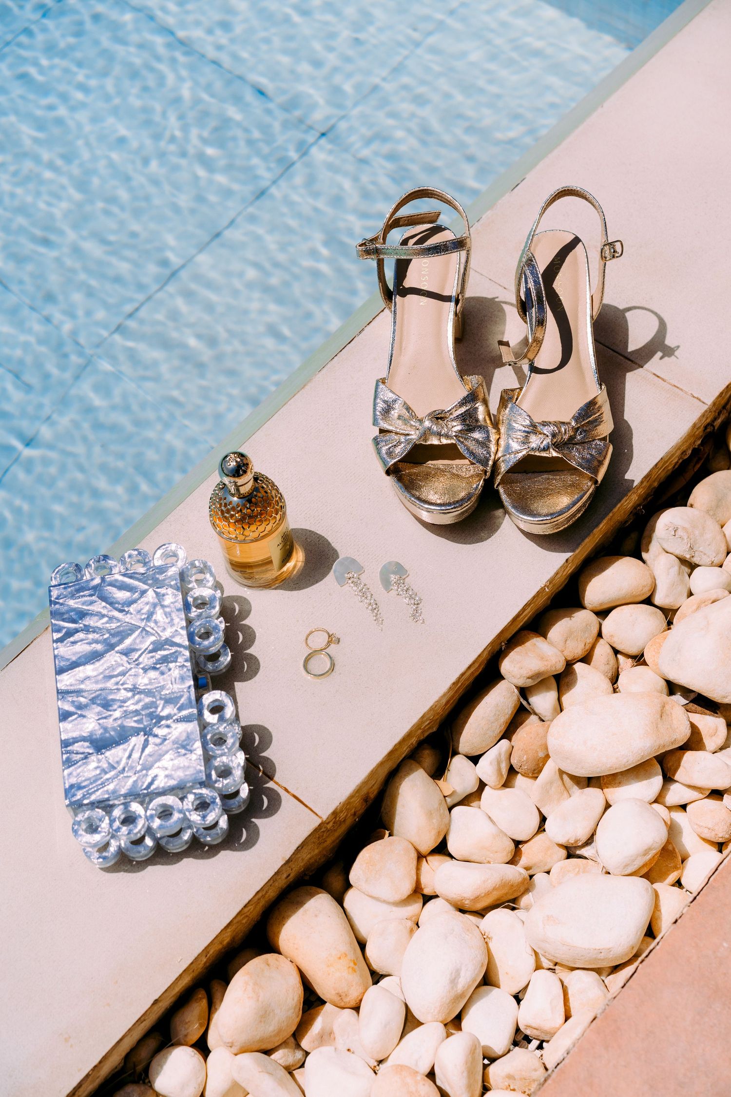 Wedding accessories including rings, perfume bottles, and white stones arranged on reflective surface.