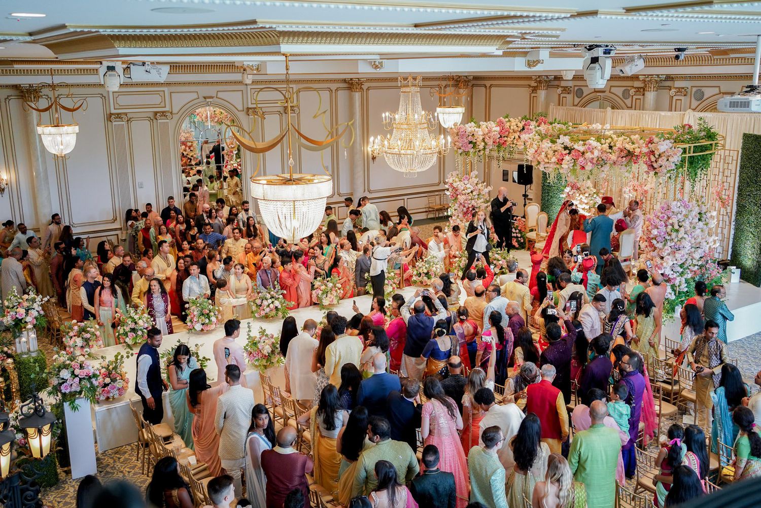 A large crowd of wedding guests dancing and celebrating on an elegant ballroom dance floor with ornate chandeliers overhead.