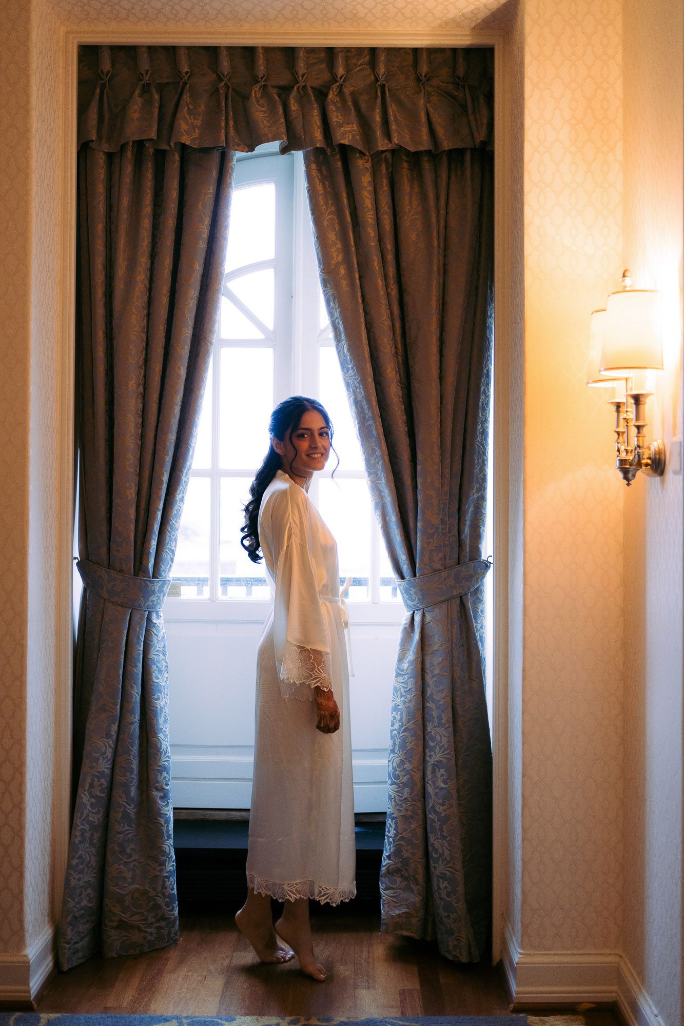 A triptych of ethereal white-clad figures standing by curtained windows in a warmly lit classical interior.