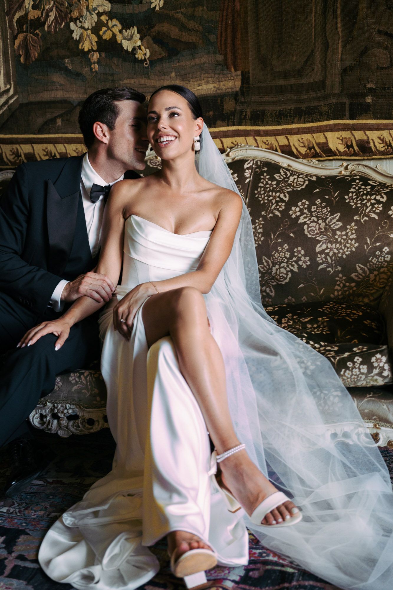 Elegant wedding couple sits intimately on vintage floral sofa with bride in white dress and flowing veil.