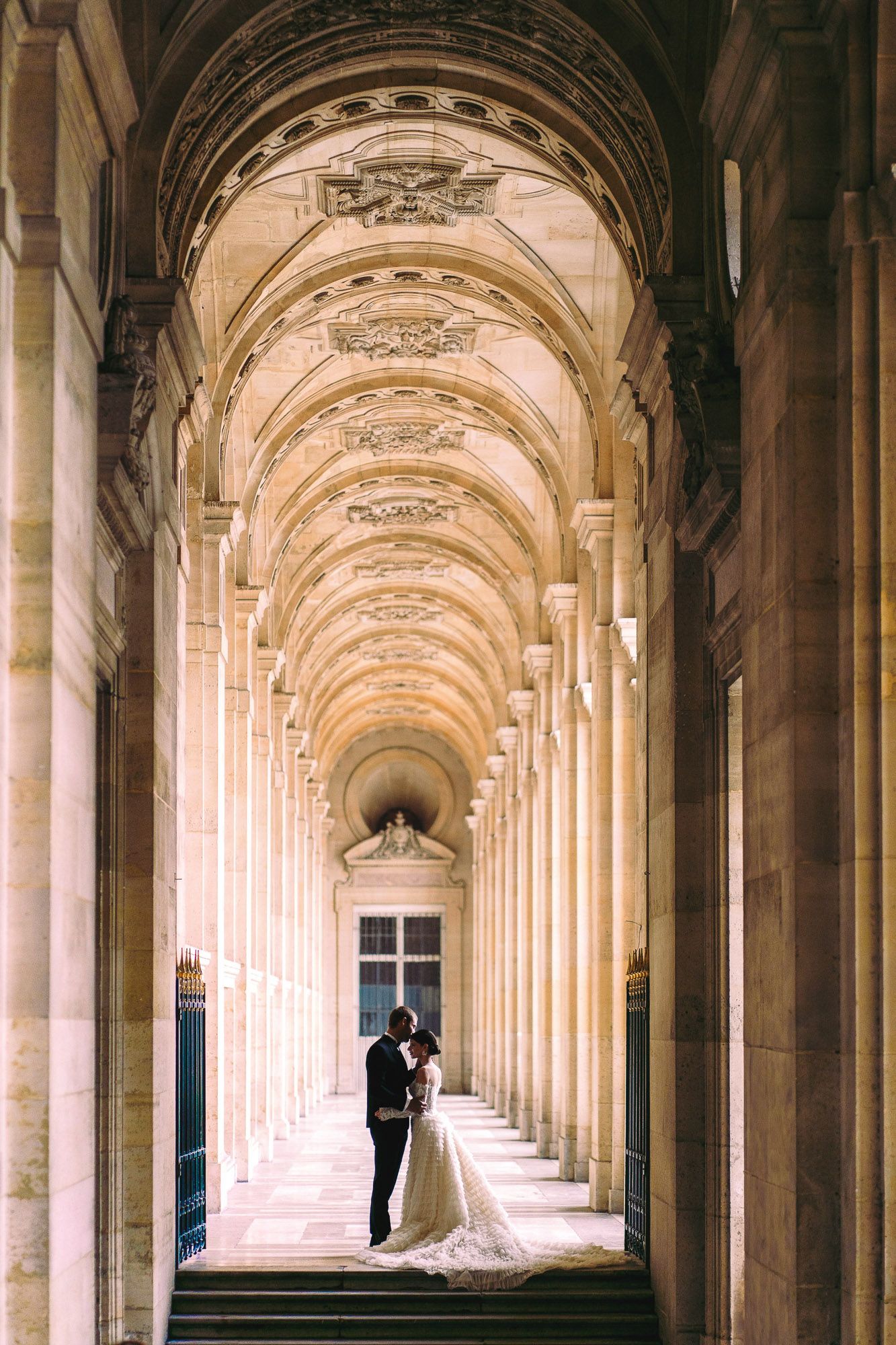 Elegant wedding photo captured in a grand colonnade hallway with arched ceilings and classical architecture.
