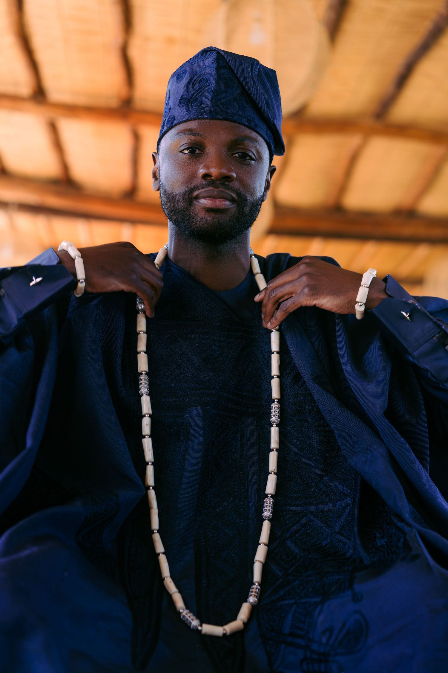 Person wearing traditional dark blue African attire with beaded necklace poses under wooden ceiling in warm light.