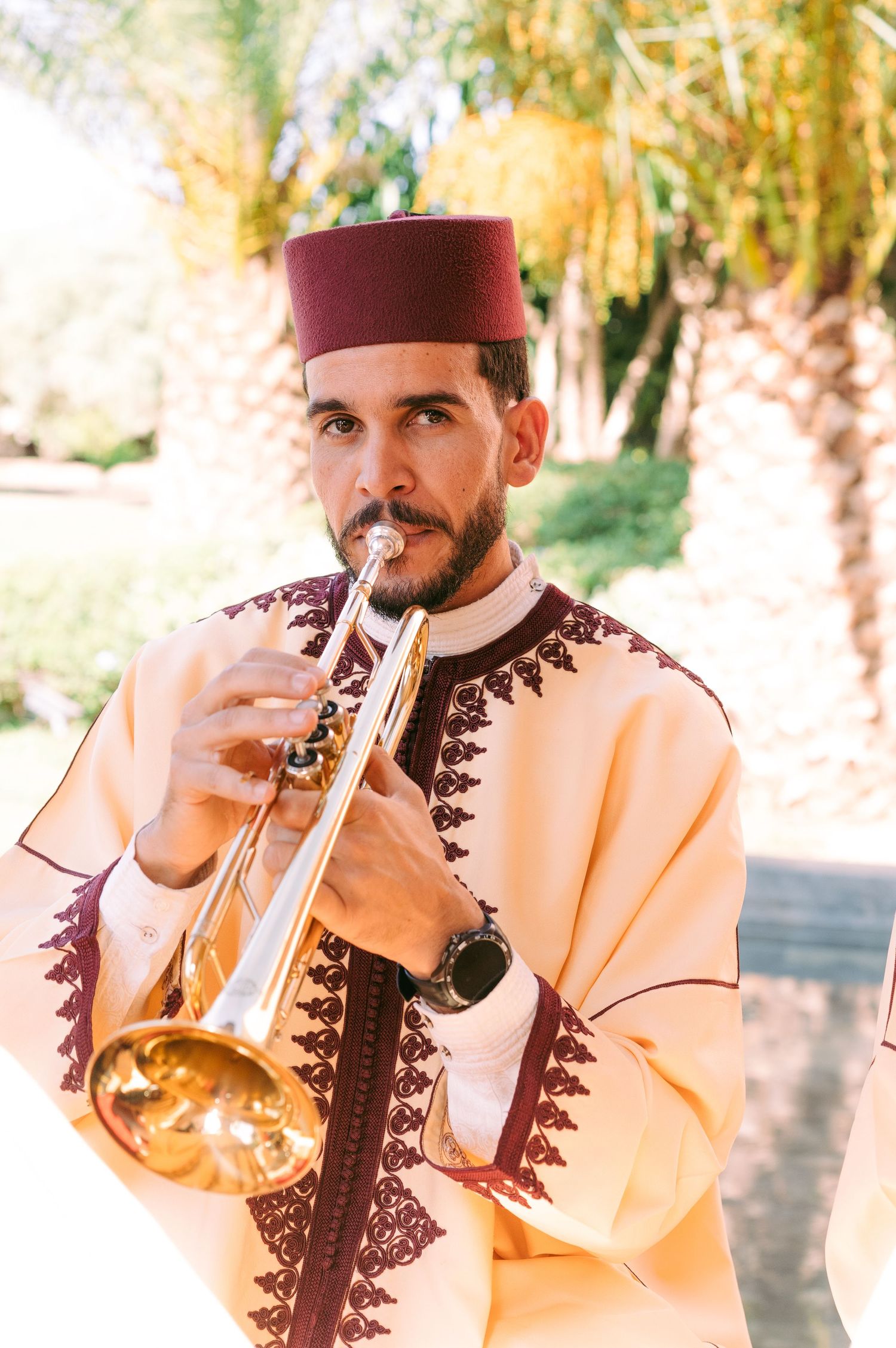 Musician in traditional Moroccan attire plays trumpet against palm tree backdrop.