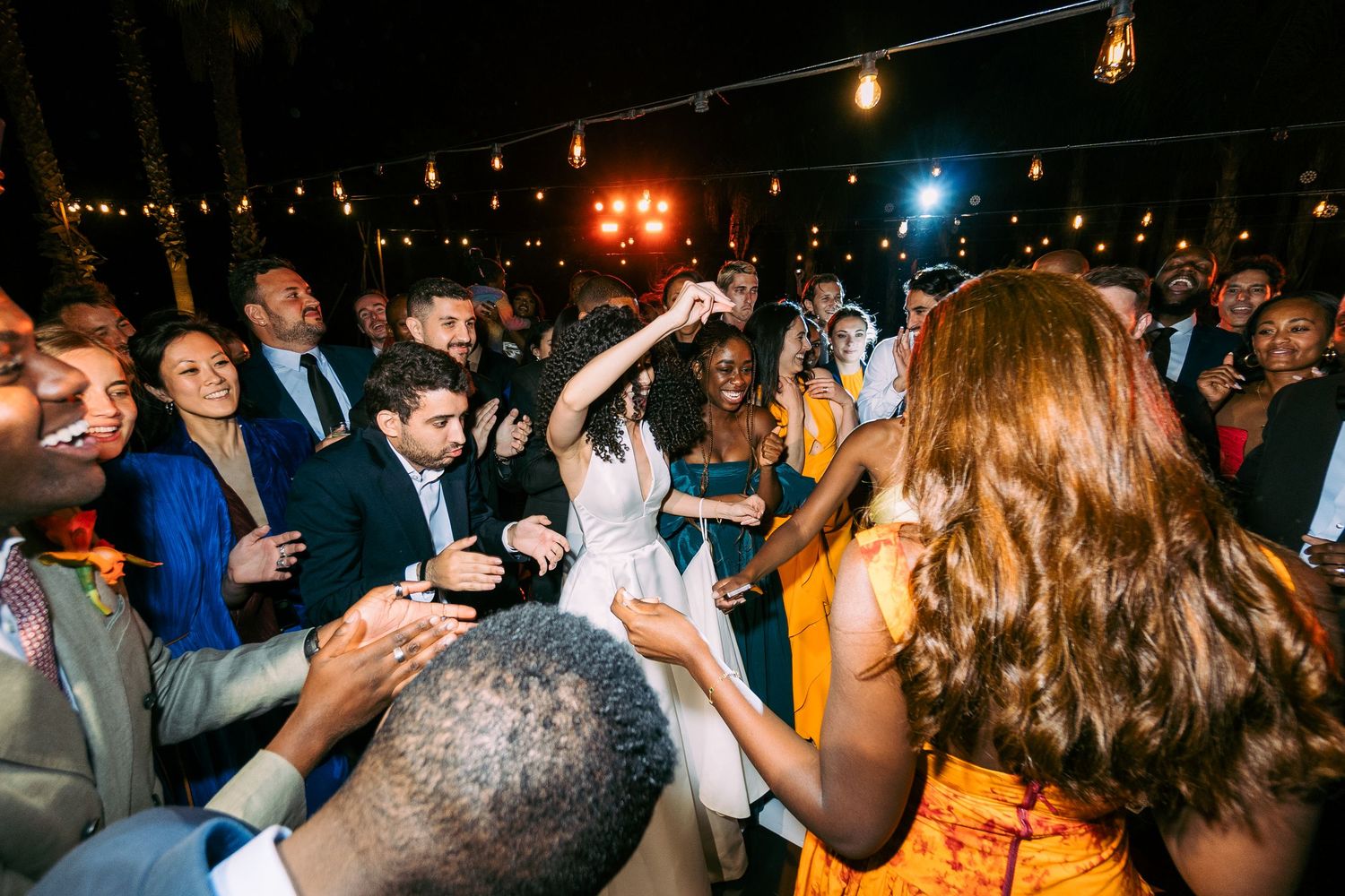 Wedding reception dance floor filled with guests celebrating under warm string lights at night.