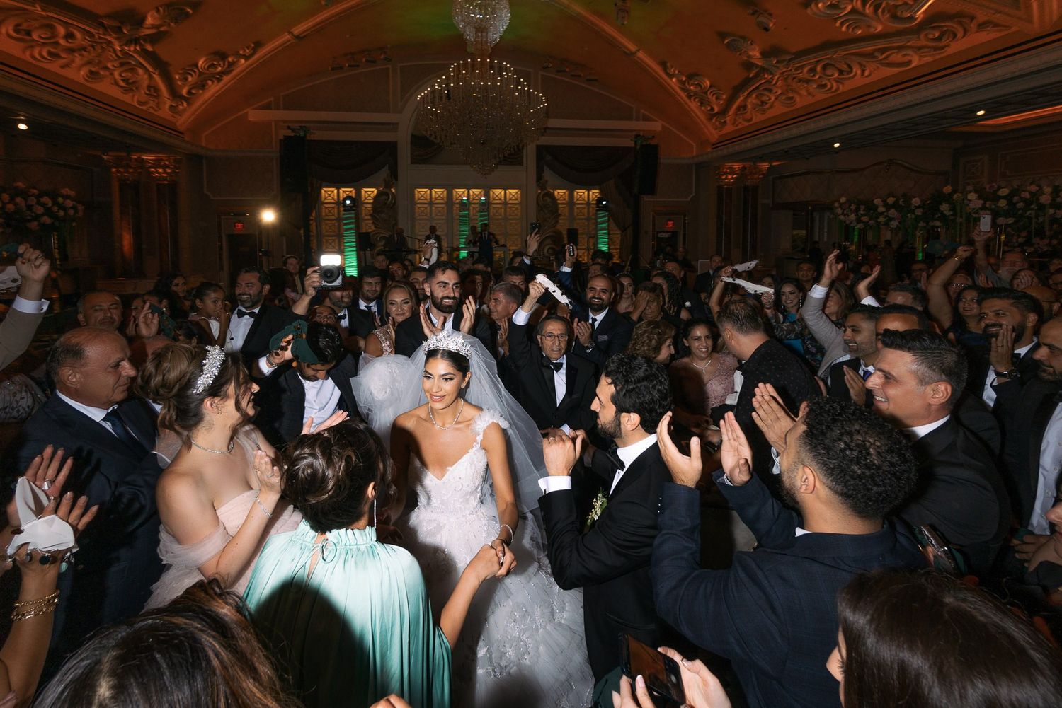 Wedding guests dance and celebrate on an ornate ballroom dance floor under elegant chandeliers during an evening reception.