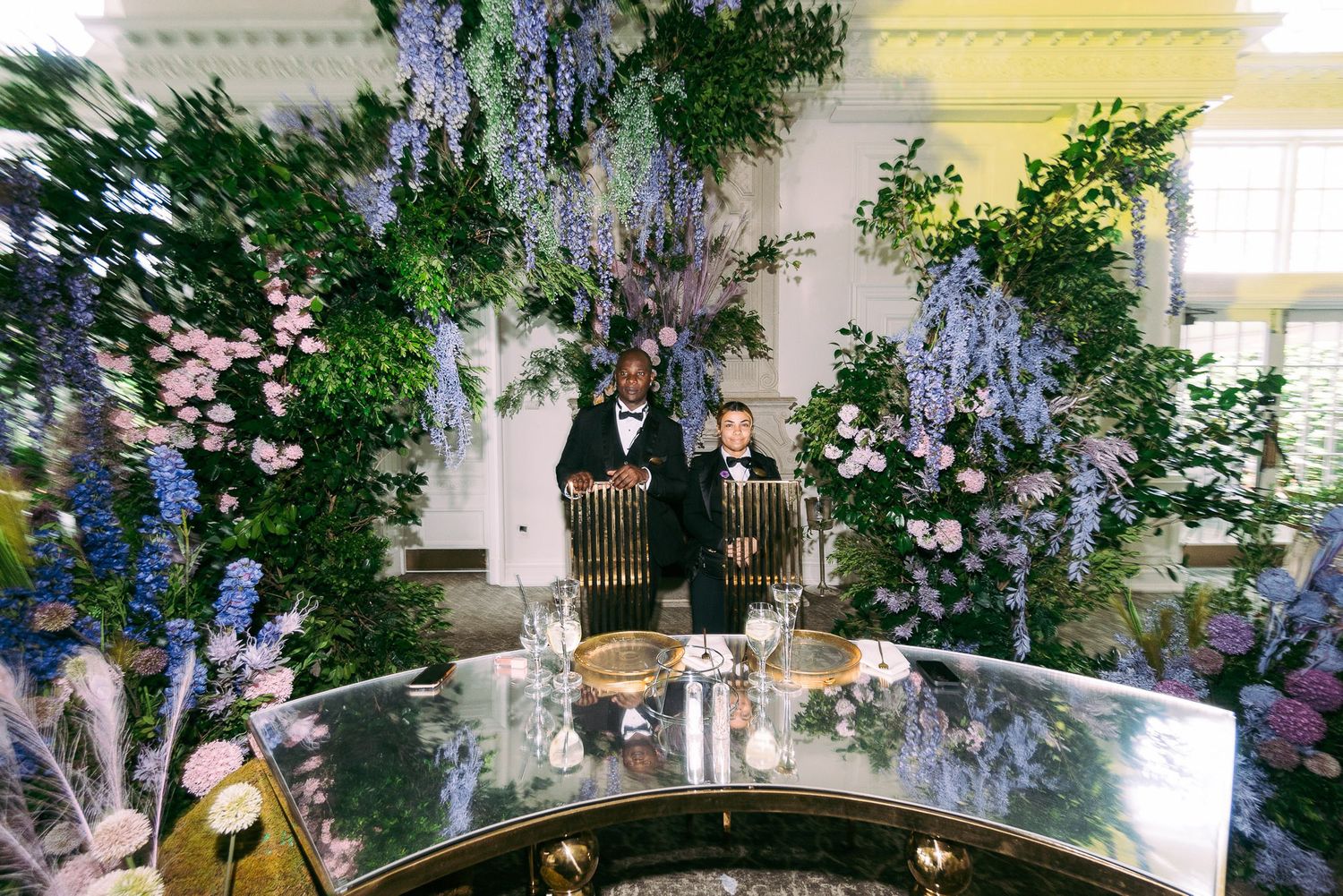 Elegant floral arrangements with wisteria and greenery create a romantic wedding reception atmosphere in a white venue.
