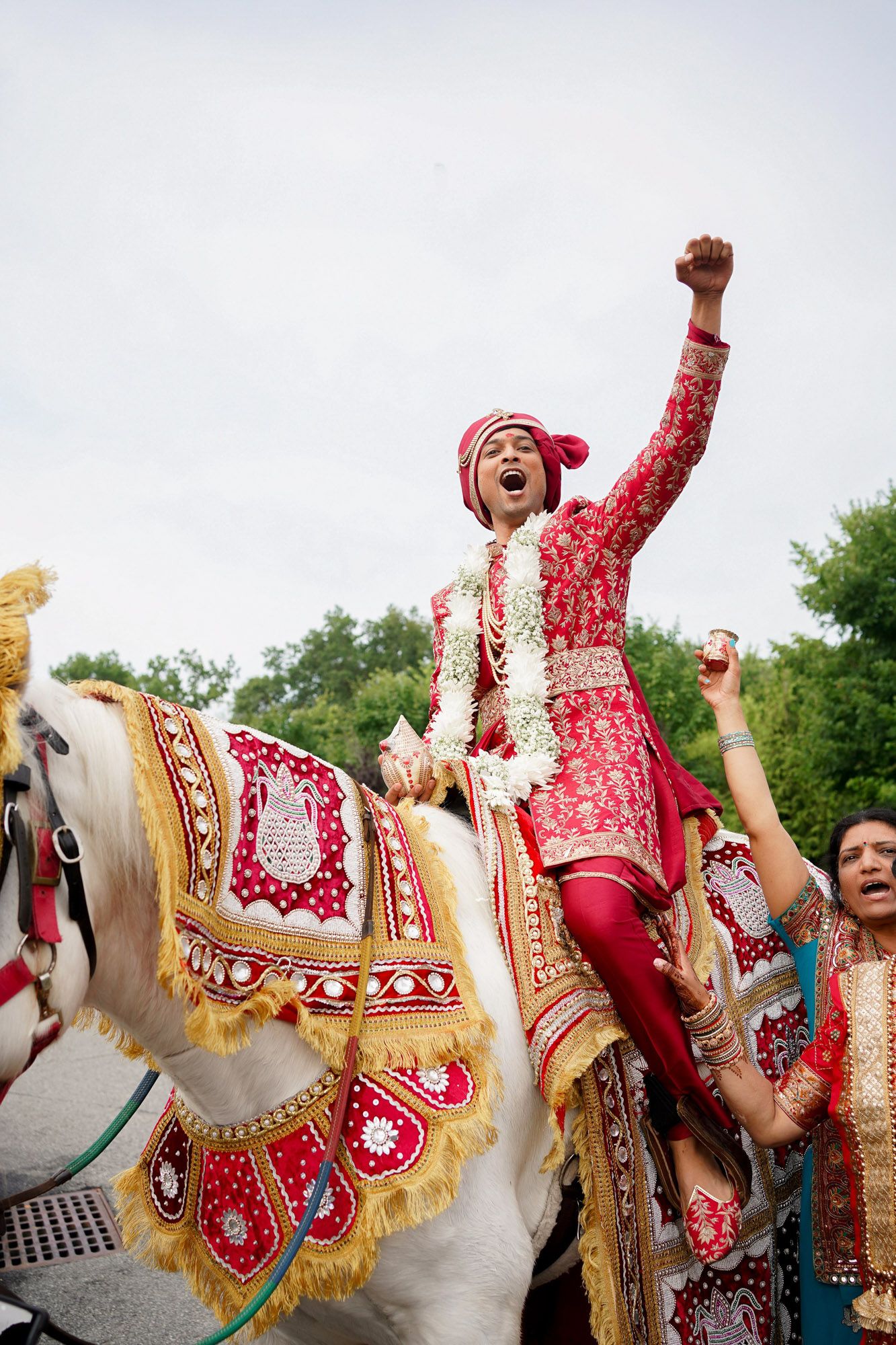 A groom in traditional red Indian wedding attire celebrates joyfully while riding a decorated white horse during a baraat ceremony.