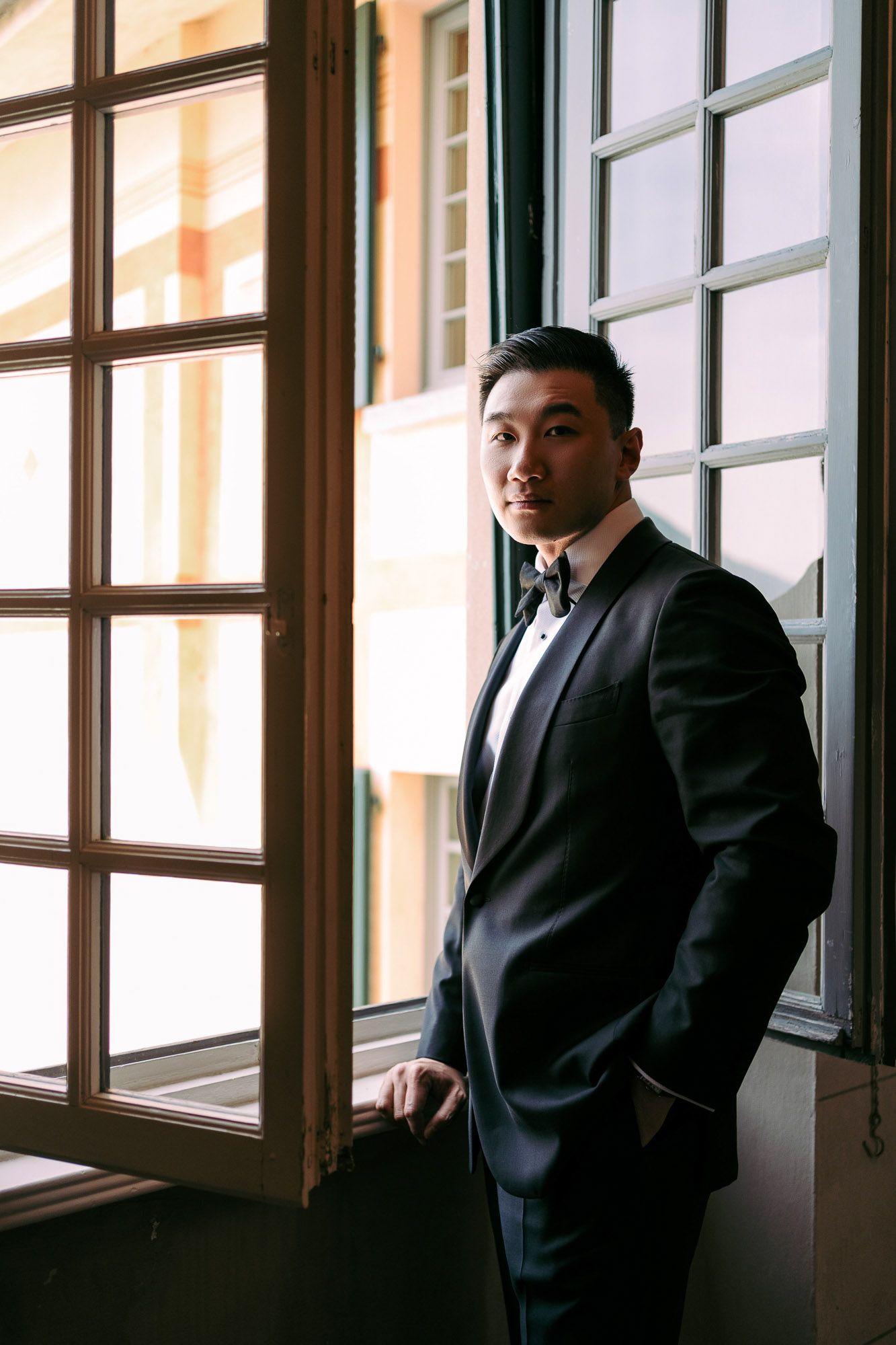 A person in a formal black tuxedo stands confidently by large windows in natural lighting.