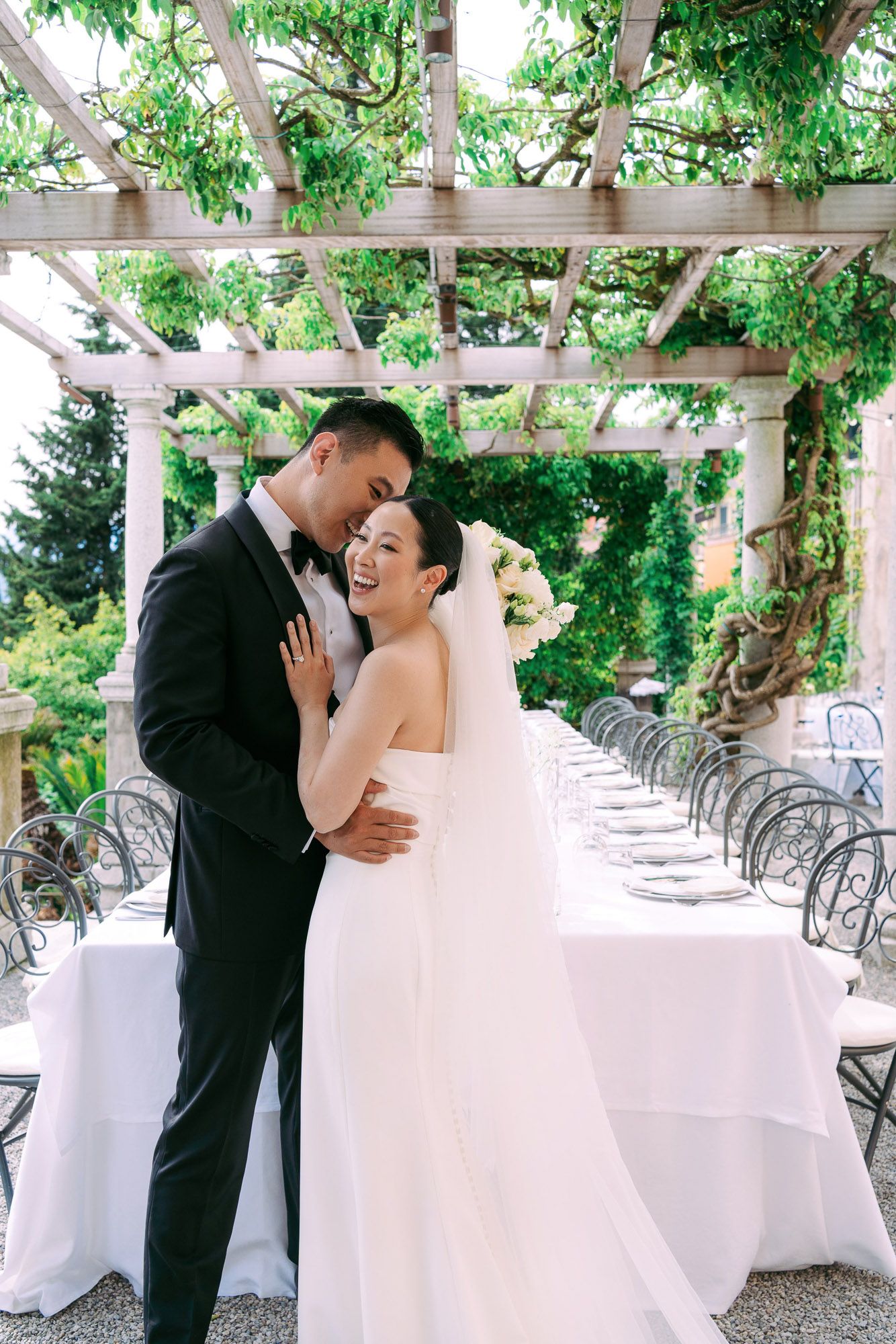 Romantic wedding couple shares tender moments under a lush garden pergola with white decor and greenery.