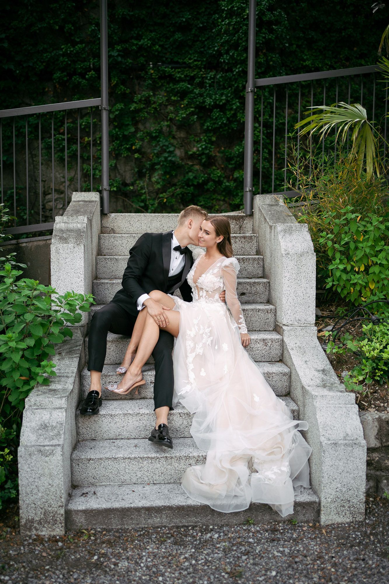A romantic wedding couple shares intimate moments while posing on stone steps surrounded by lush greenery.